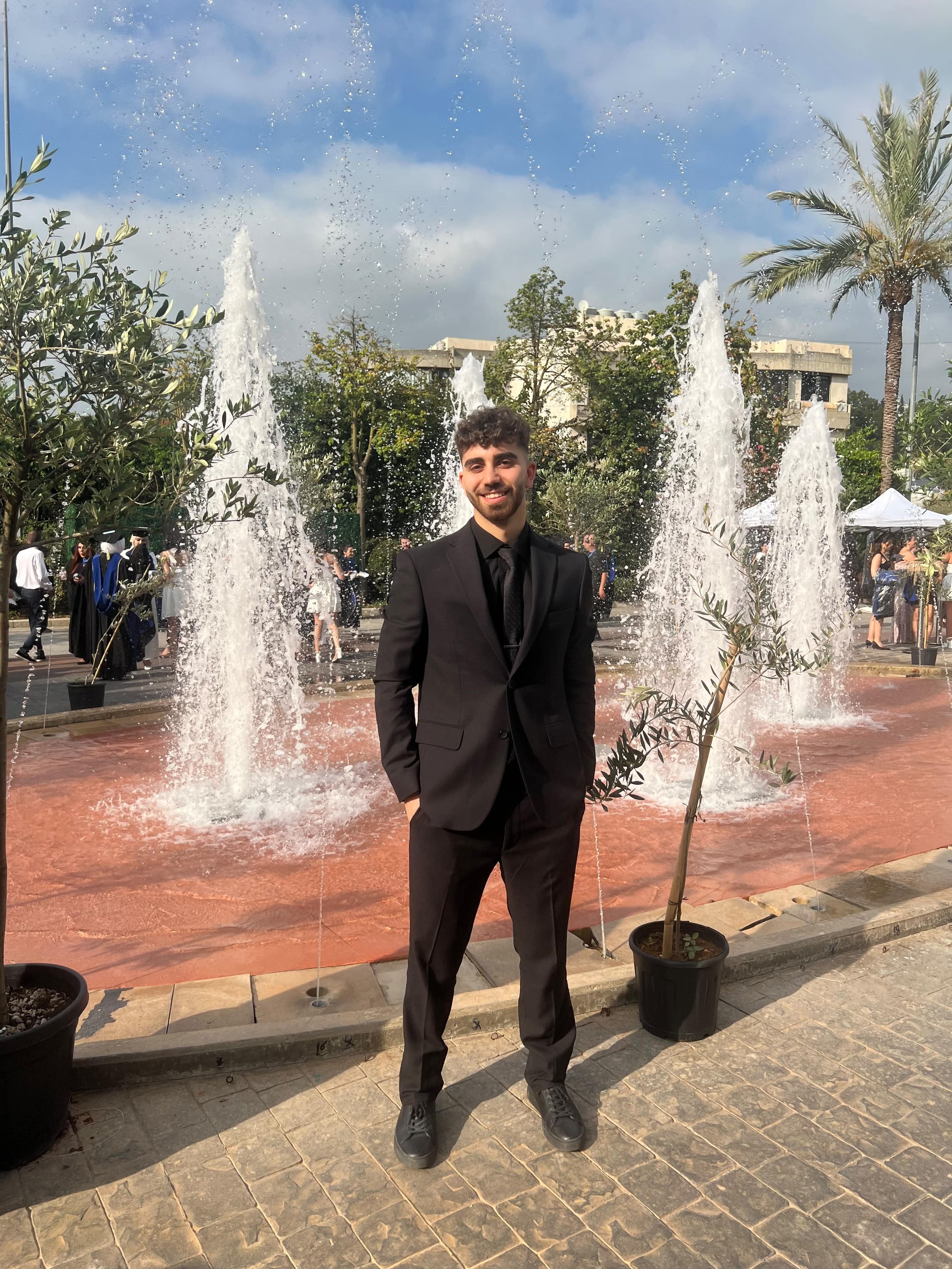 A young man in a black suit and black shoes standing in front of a fountain with water jets shooting upward. There are potted plants, trees, and people in the background at an outdoor event or celebration under partly cloudy skies.