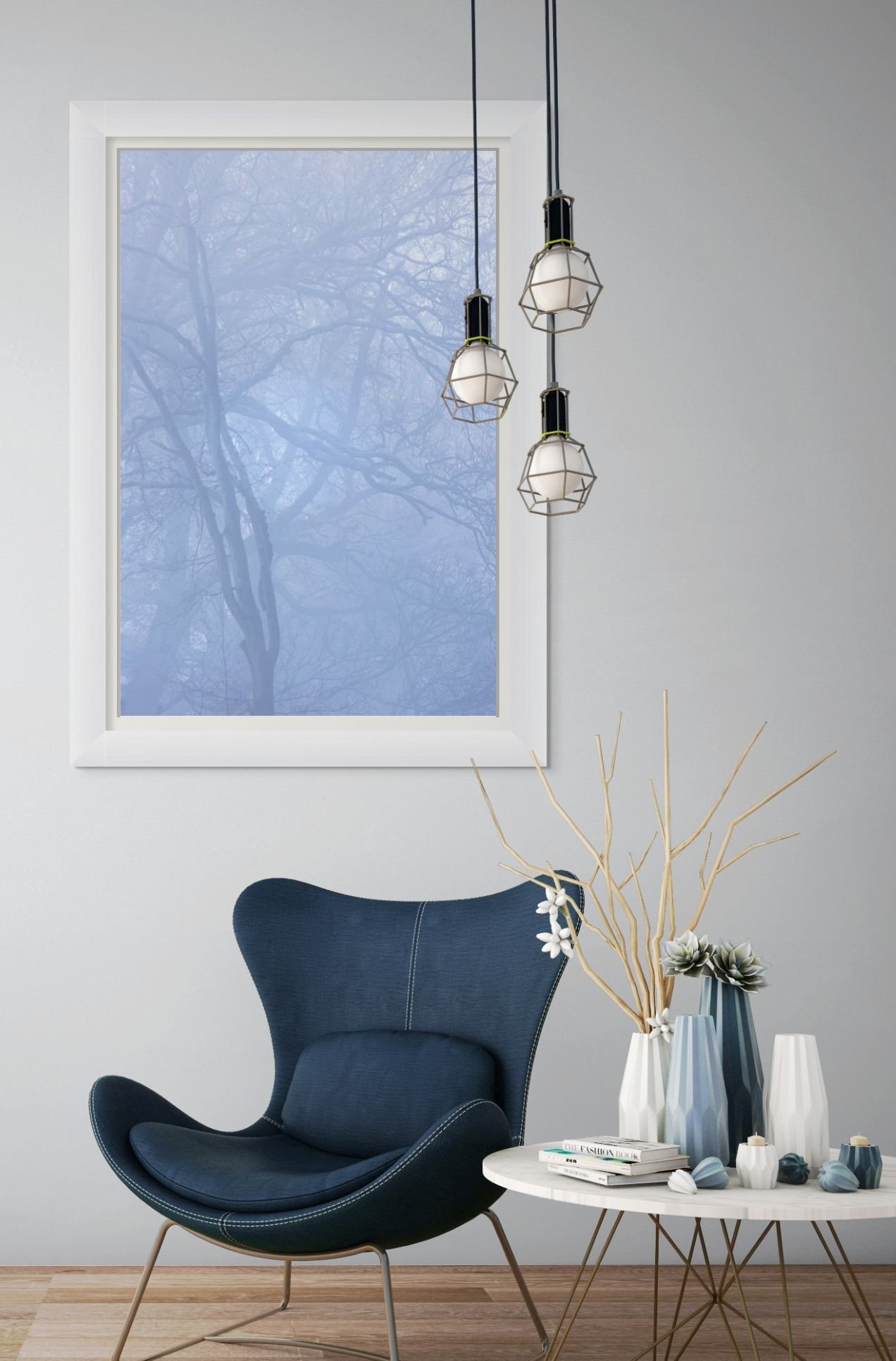 Pale blue fine art photograph of misty tree branches with a white frame in a living room with a denim blue chair, table ornaments and three modern ceiling lights