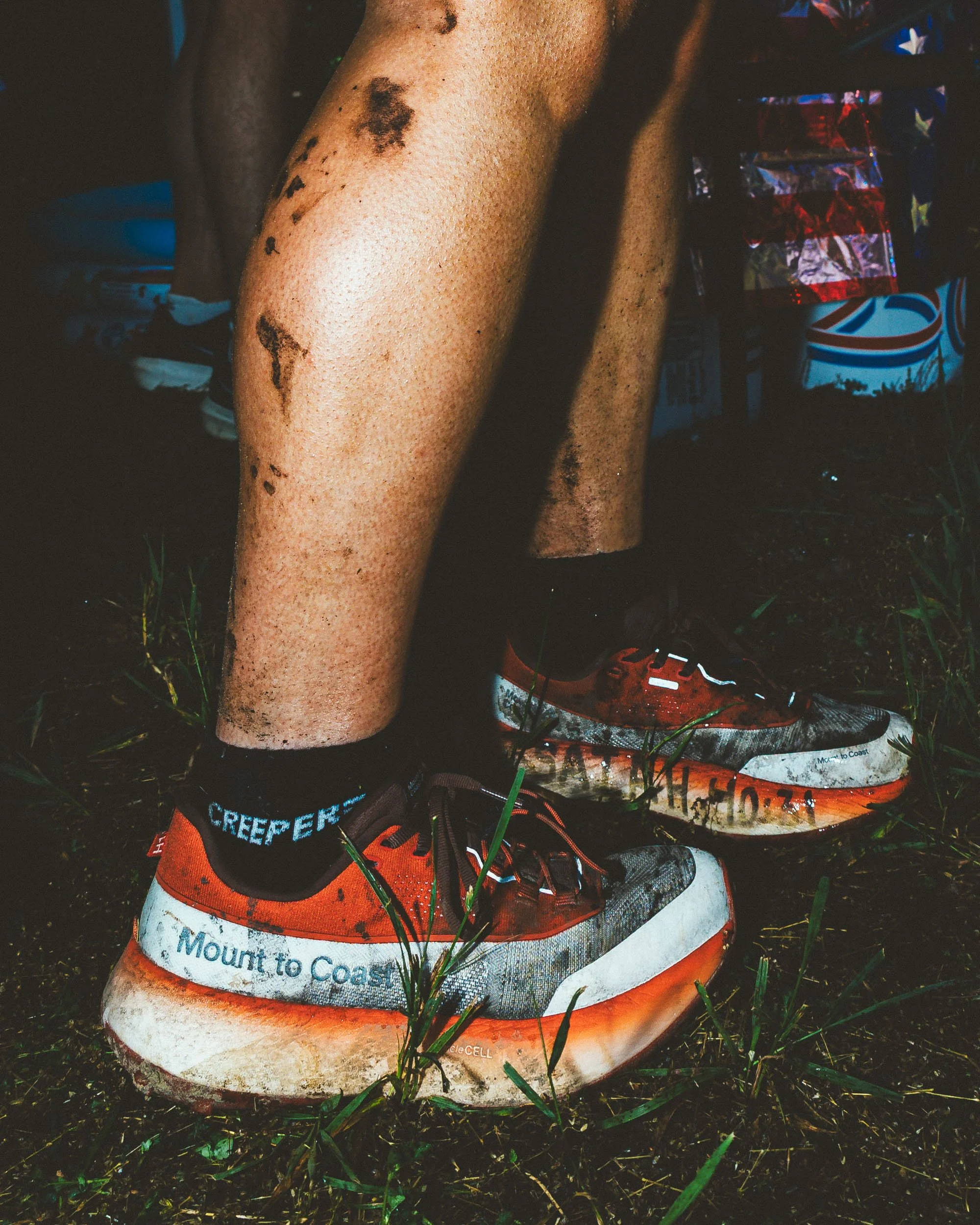 Close-up of a muddy trail runner's shoe and calf, with some tattoos on the leg, on grass and dirt ground. The shoe has a race bib attached with the words 'Mount to Coast' and 'CREEPER' on the sock.