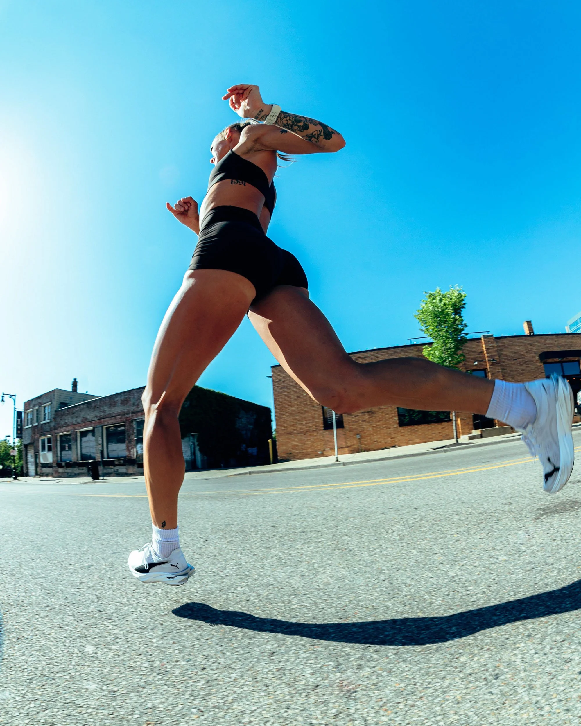 A person with tattoos is performing a handstand on the street in front of brick buildings, wearing athletic clothing, with a clear blue sky overhead.