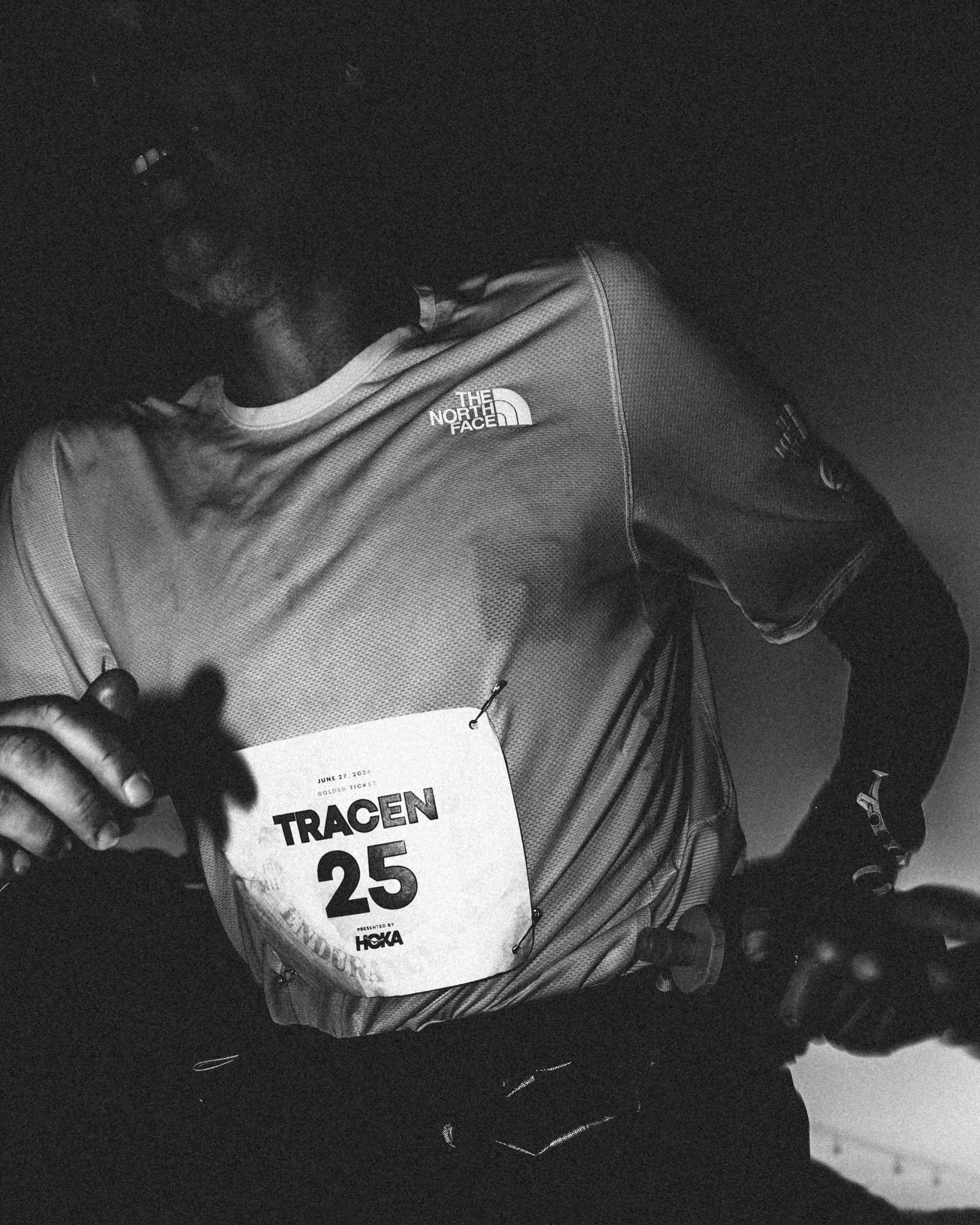 Black and white photo of a person holding a race bib with the number 25, the person is wearing a North Face jacket and appears to be at a running event.