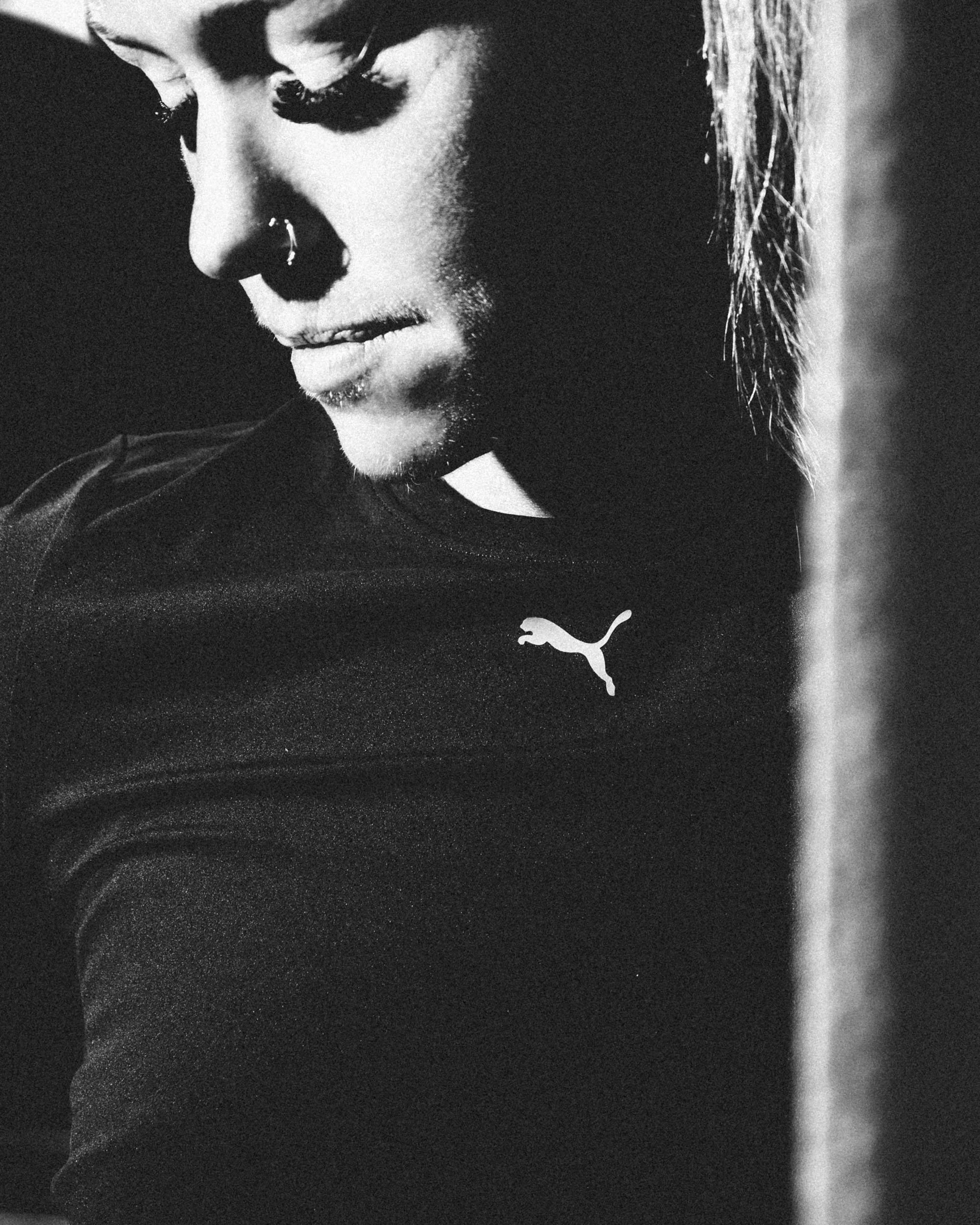 Close-up black and white photograph of a woman's face, with her eyes closed and head slightly tilted downward, wearing a dark Puma sports shirt.