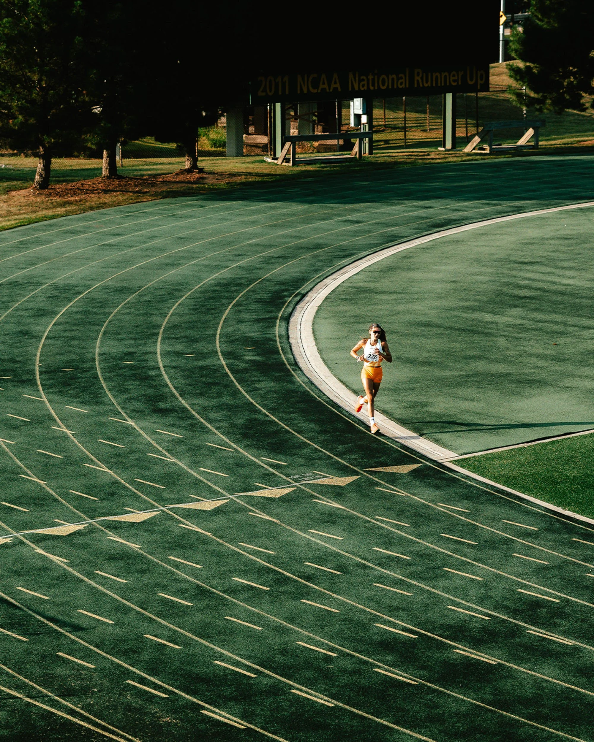 A female runner wearing a bib number 228 running on a green outdoor track with lanes marked in white, surrounded by trees and benches, with a sign in the background indicating the 2011 NCAA National Runner Up.