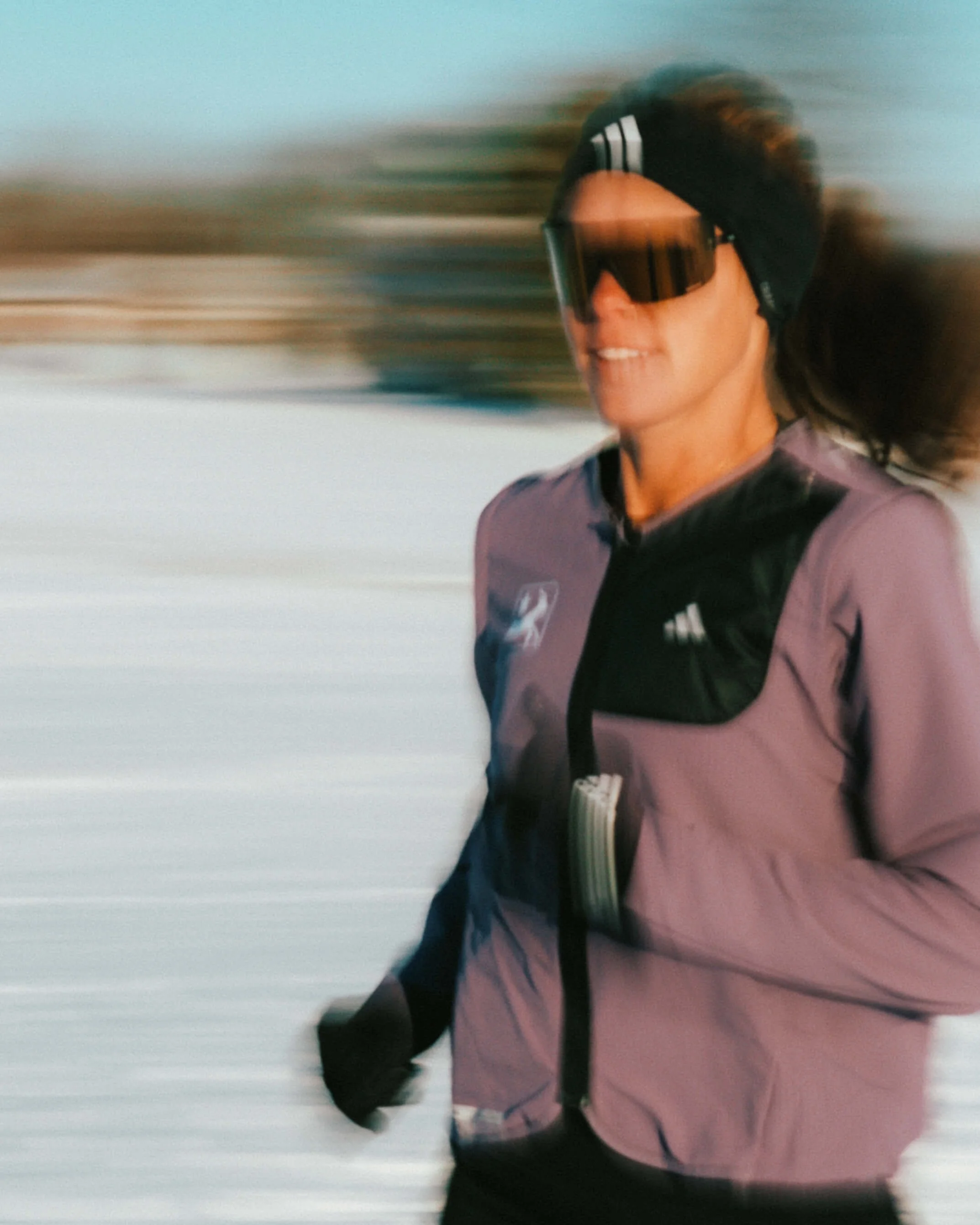 A woman running outdoors, wearing sunglasses, a pink and black jacket, and a black beanie, with a blurred background suggesting motion.