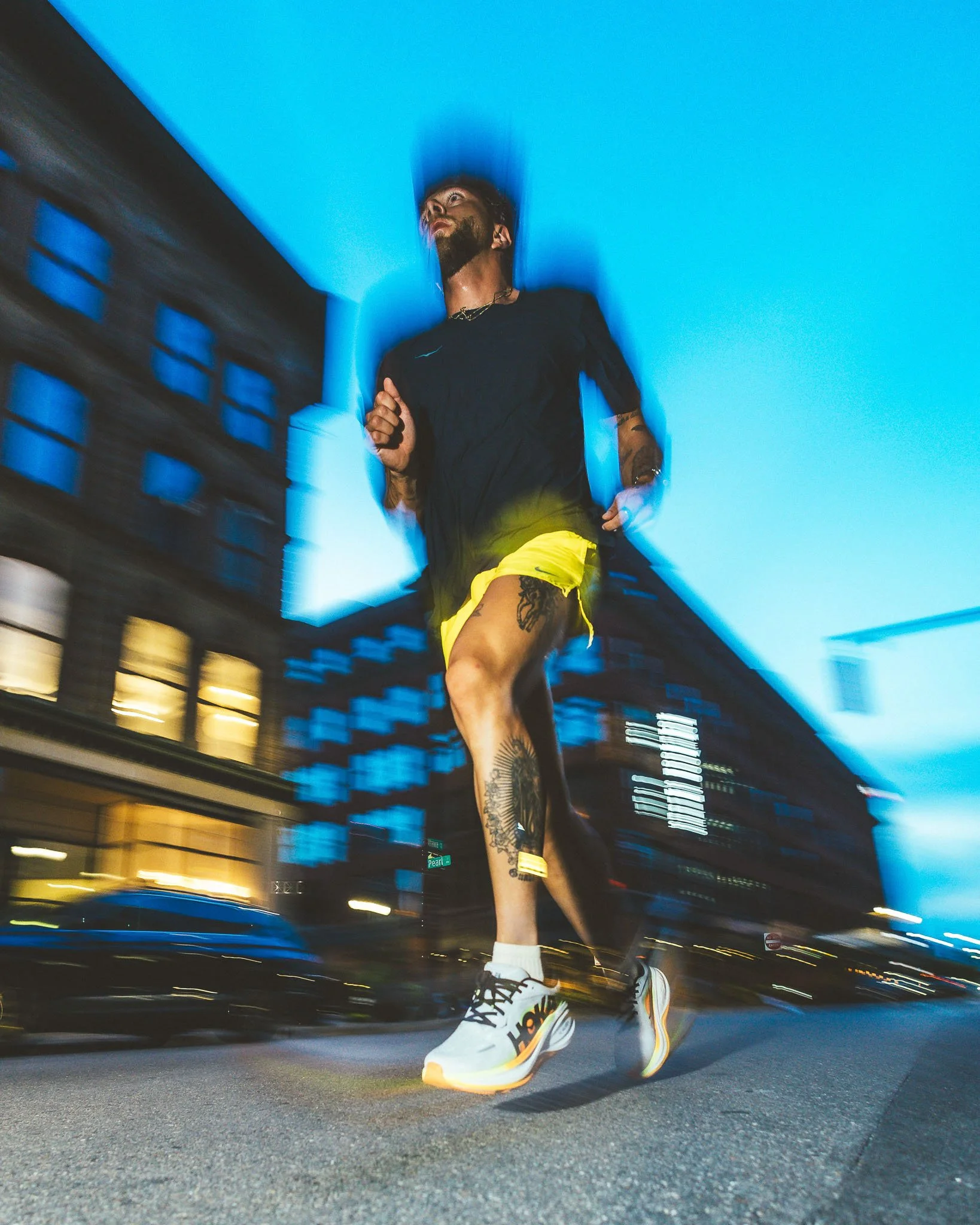 A man running on a city street at night, viewed from a low angle, blurred lights and a modern building in the background, wearing a black t-shirt, yellow shorts, white sneakers, and sporting tattoos.