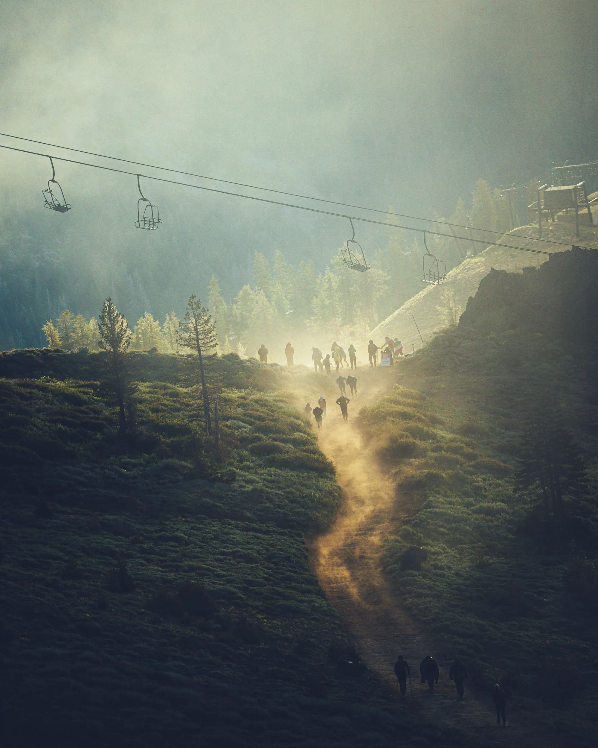 Mountains with mist, cable cars suspended in the air, a dirt trail with hikers walking, and a small building on the hillside.