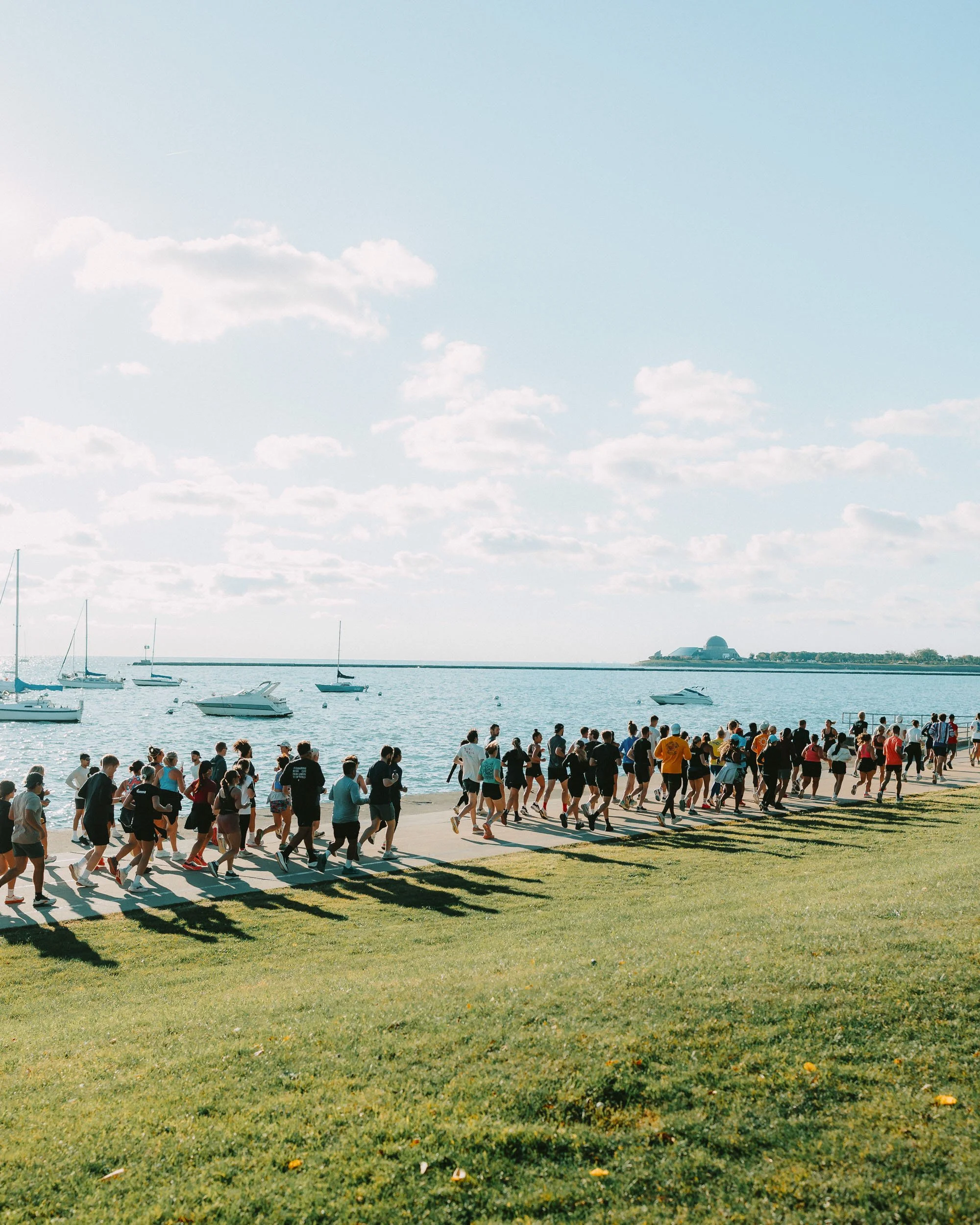 A large group of people participating in a running event along a waterfront, with boats in the water and a building with a dome in the background.