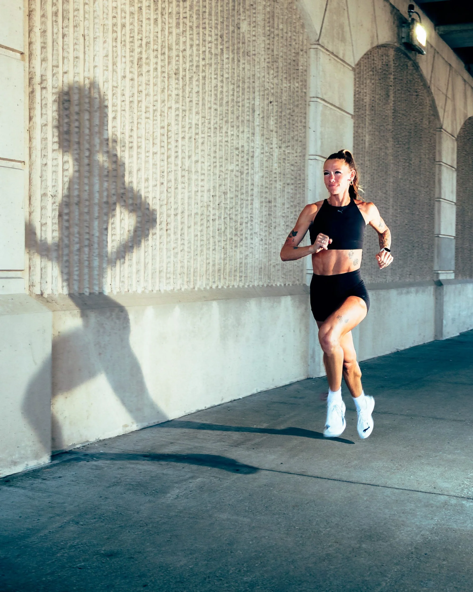 A woman in black athletic wear running outdoors under a bridge, with a shadow of her right arm on the wall.