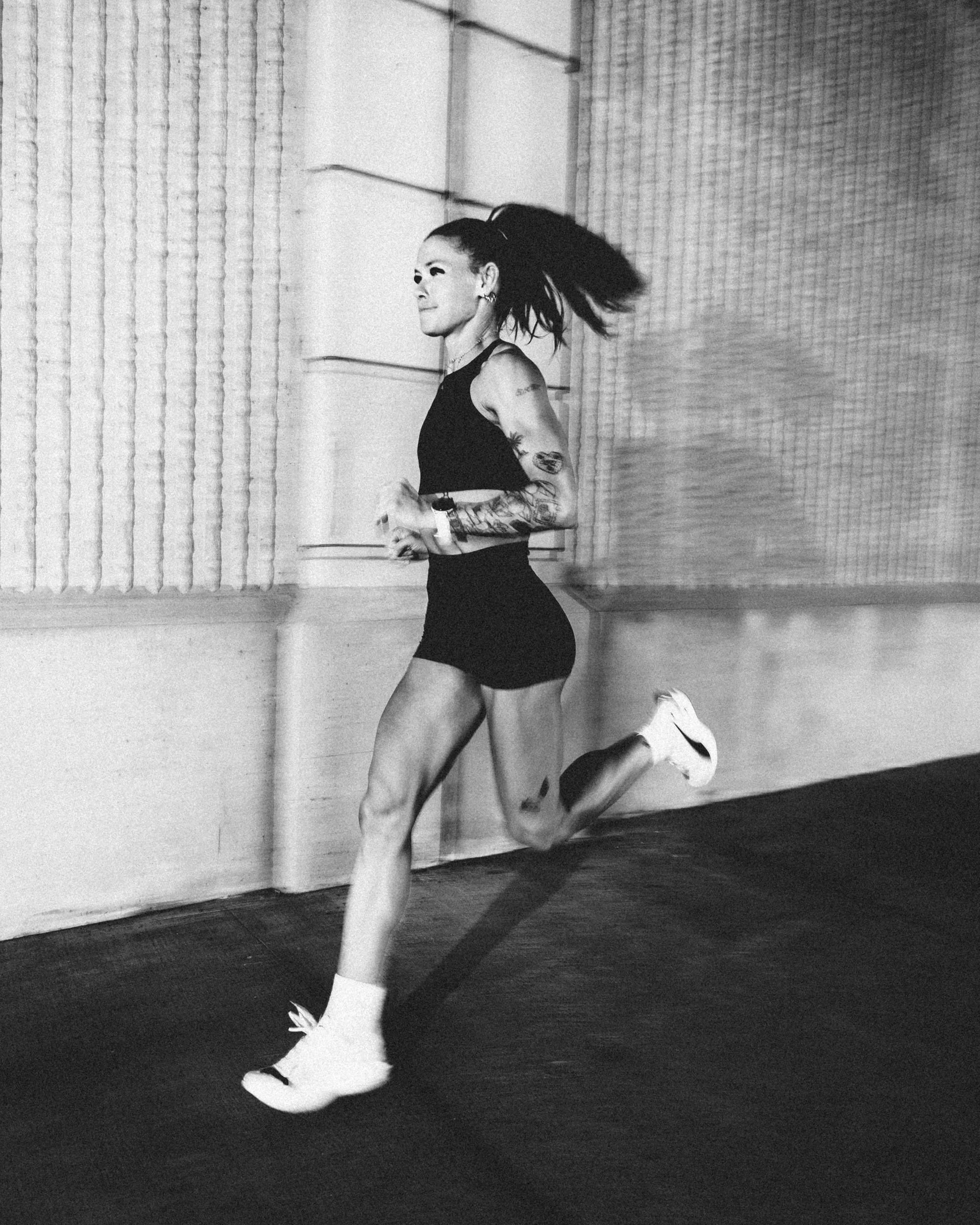 A woman with tattoos and long dark hair tied in a ponytail is running indoors on a dark wooden floor, wearing athletic clothing and white sneakers.