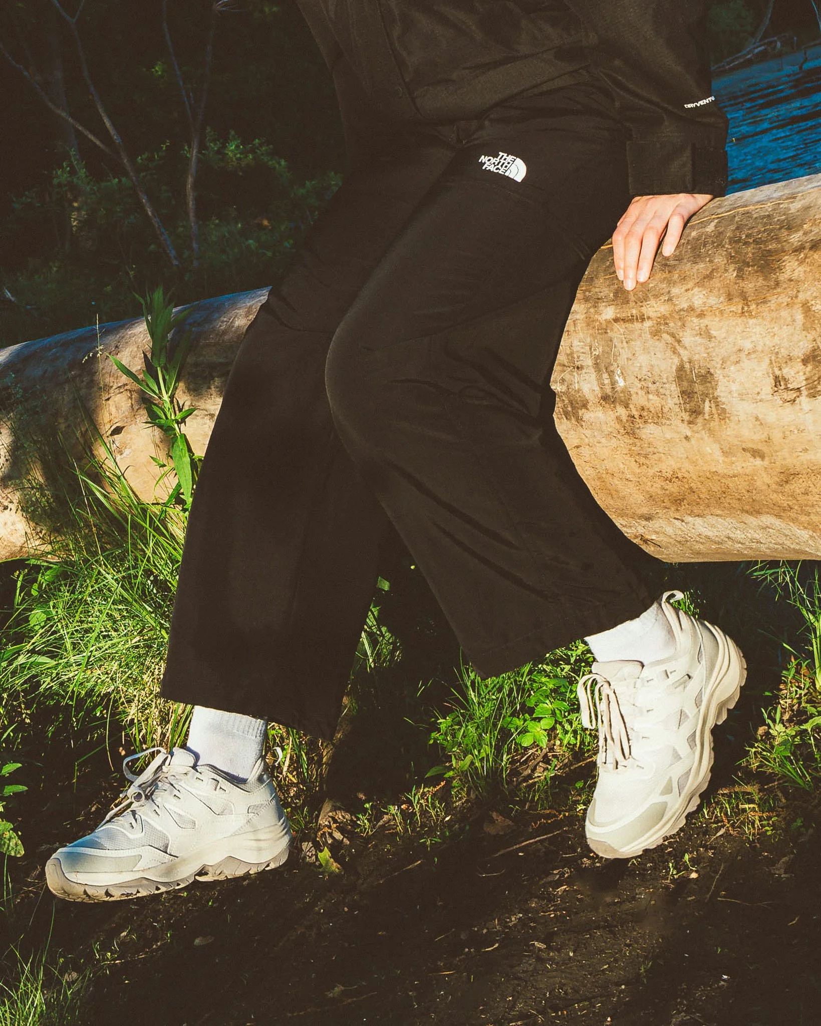 Close-up of a person's lower body, wearing black pants with The North Face logo, white sneakers, standing on dark soil with green grass and small plants nearby, in an outdoor natural setting.