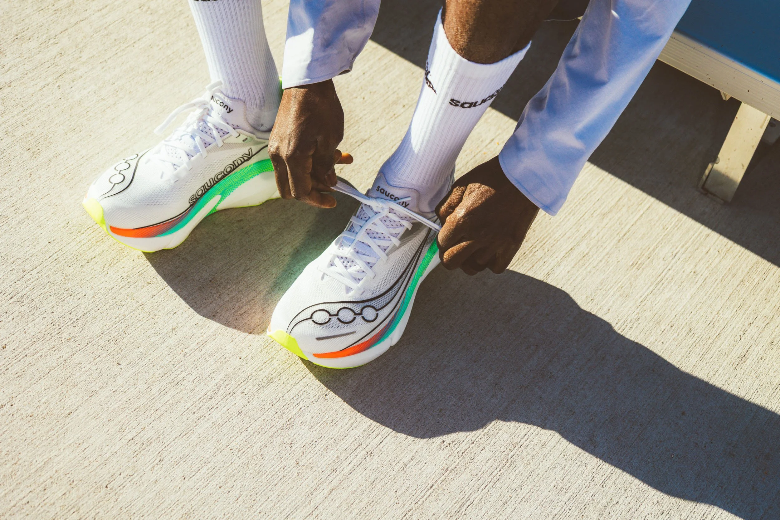 Person tying shoelaces on white athletic shoes with colorful accents, sitting on a bench outdoors.