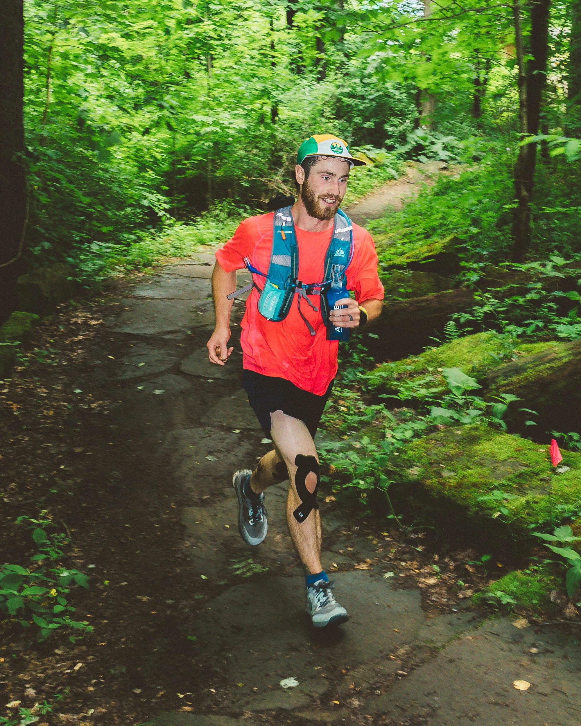 A man running on a trail in a lush, green forest. He is wearing a colorful cap, a bright orange T-shirt, a hydration pack, and running shoes. The trail is surrounded by dense foliage and trees.