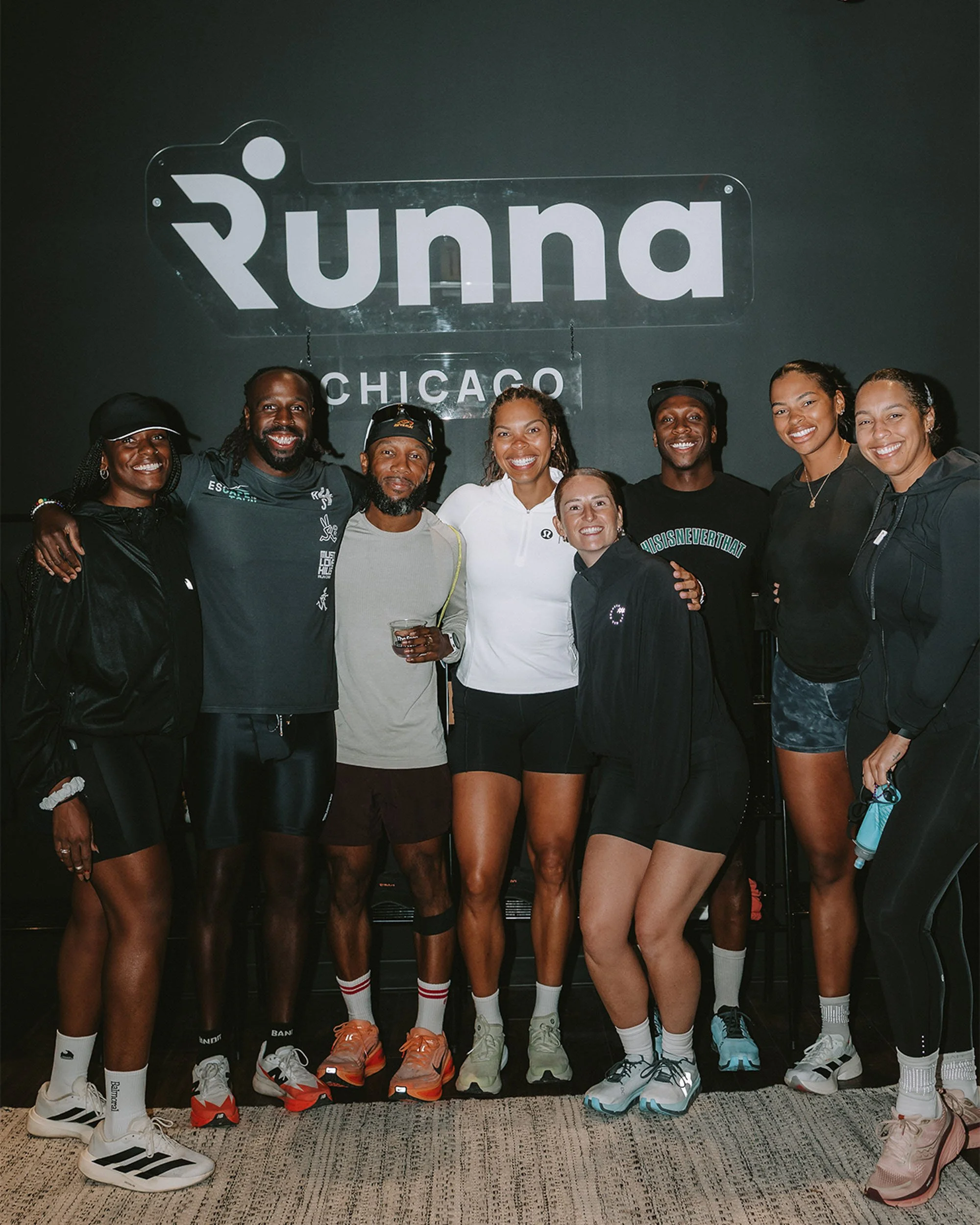Group of eight diverse athletes in running gear smiling and posing together in front of a dark wall with a sign that reads 'Running Chicago'