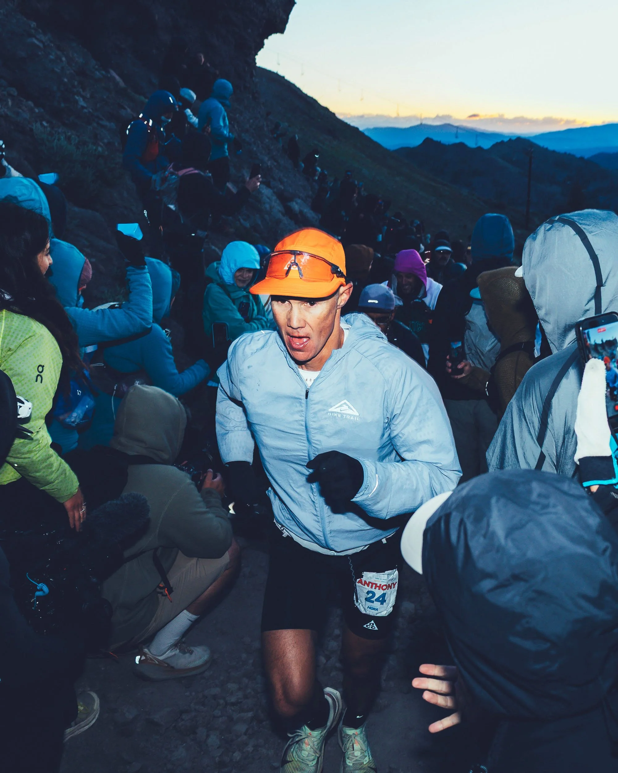 A group of runners dressed in athletic gear and jackets, some with hooded caps or hats, are participating in an outdoor race on a rocky trail during dawn or dusk with mountains and a faint sunset in the background.
