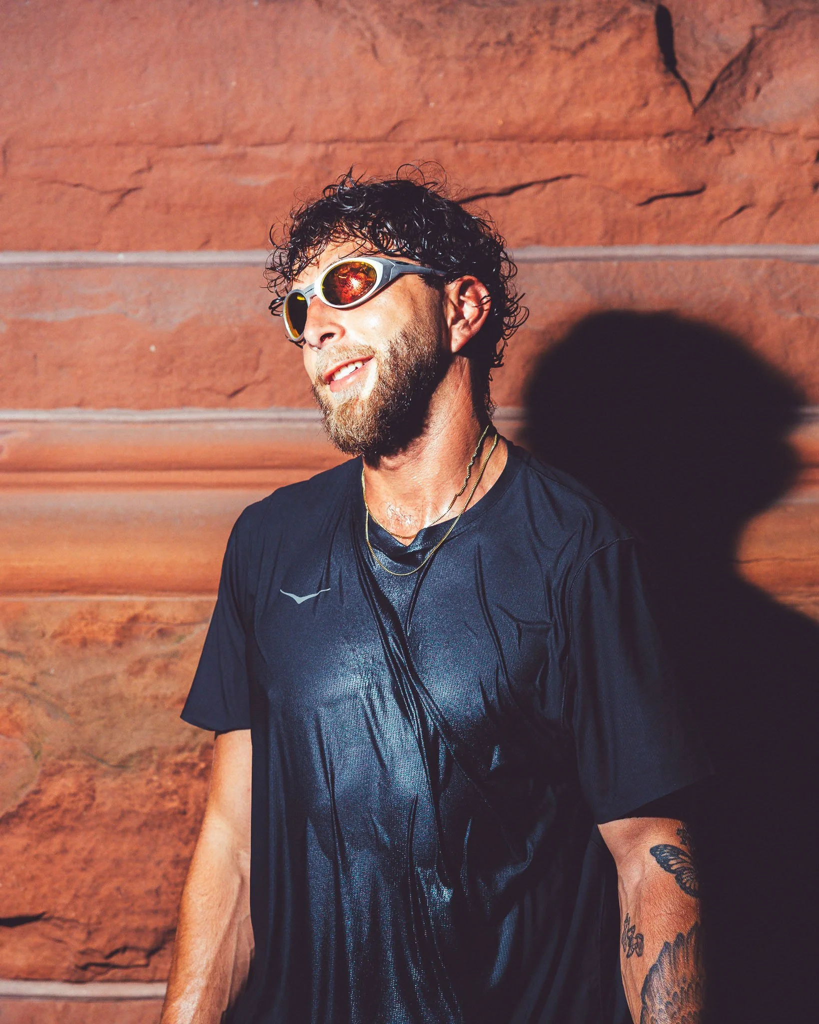 A man with curly dark hair and a beard wearing reflective sunglasses, a black shirt, gold necklaces, and tattoos on his arm, standing in front of a reddish-brown wall with horizontal lines.