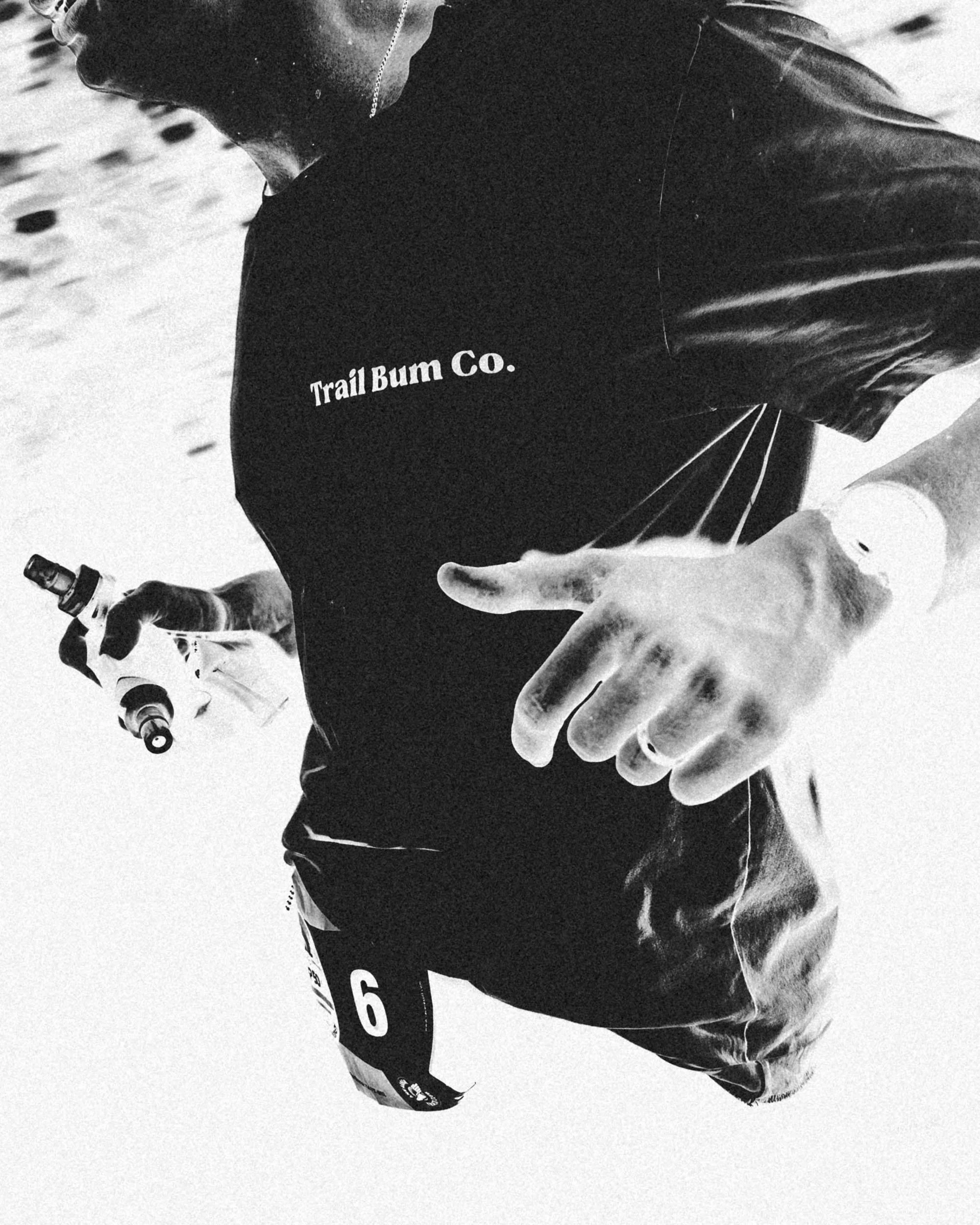 Black and white photo of a person wearing a dark jacket with the words 'Trail Bum Co.' on it, holding a small bottle, with a large hand in the foreground.