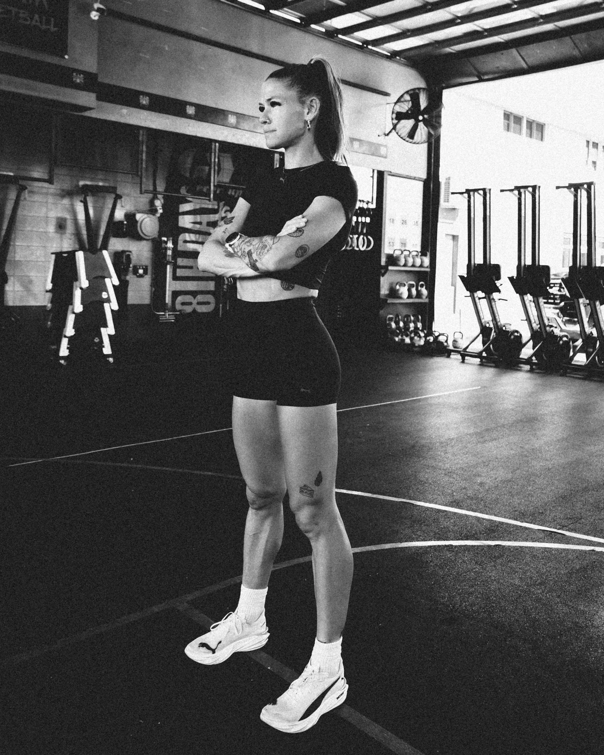 A woman with tattoos standing in a gym, wearing sportswear, crossing her arms, and looking to the side.