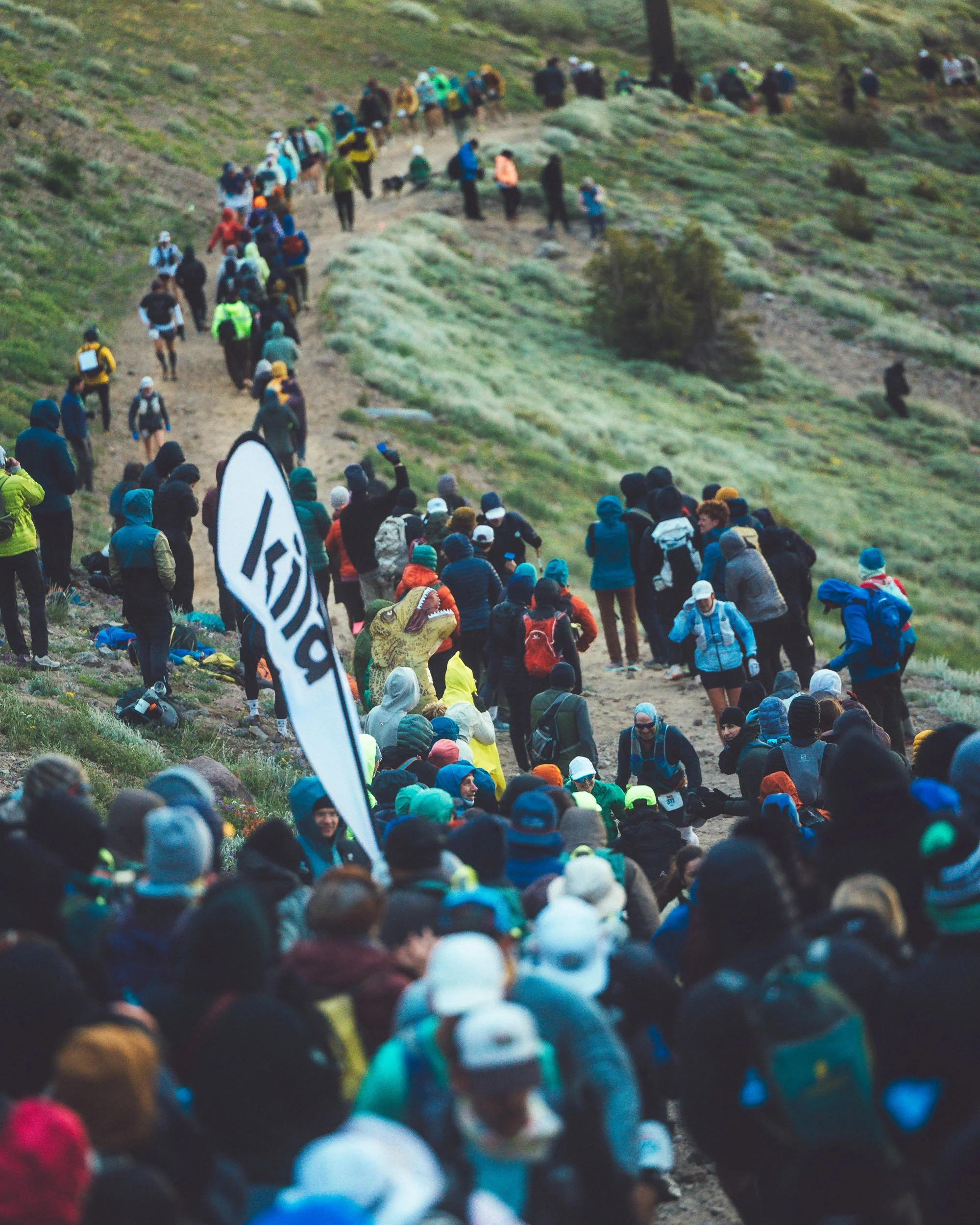 A large group of people hiking on a mountain trail, many wearing colorful outdoor jackets and hats, with some carrying backpacks and others taking photos, with grassy terrain and hills in the background.
