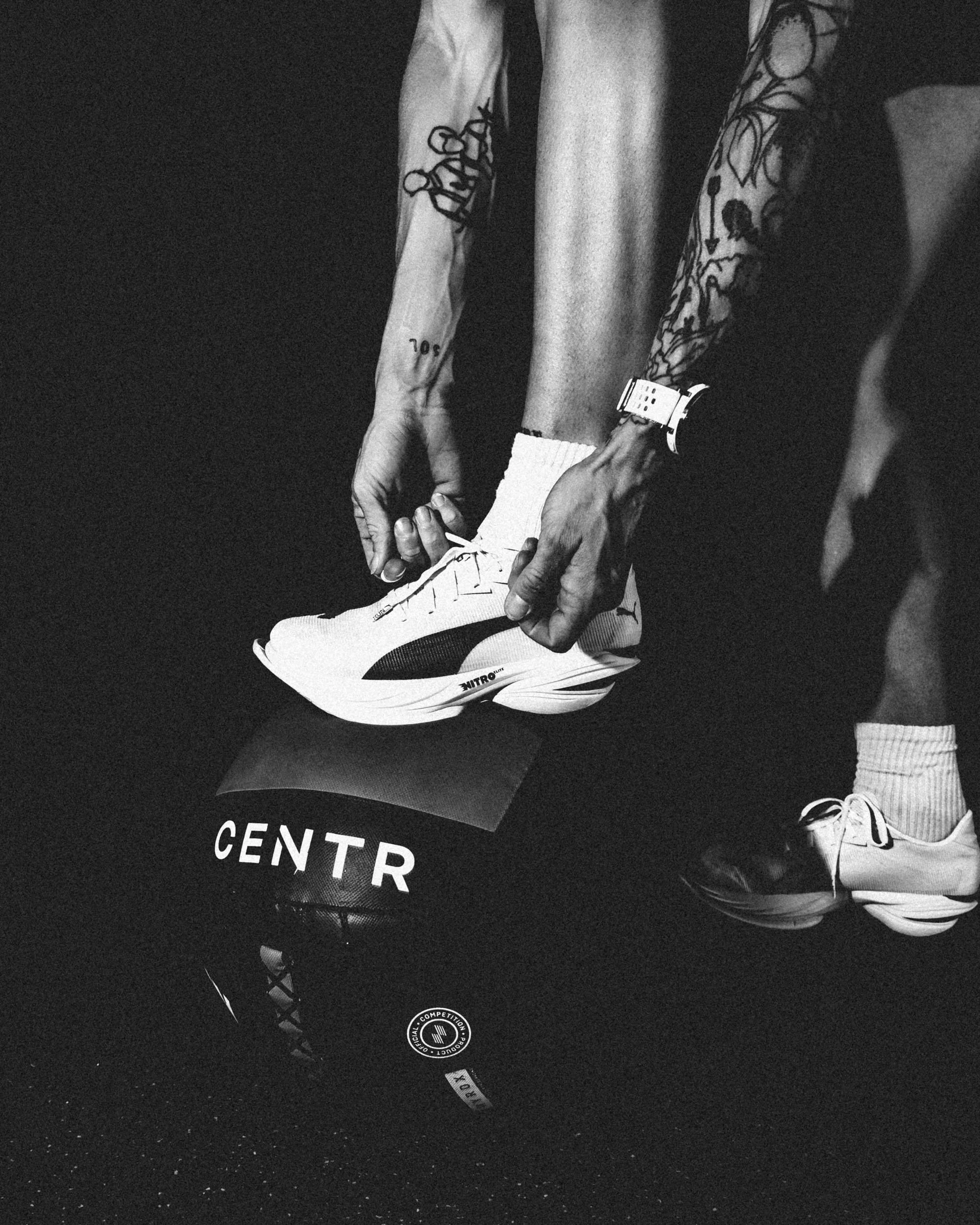 A person adjusting white athletic shoes with black accents while wearing white socks, a black apparel with a circular logo, and having tattoos on their arms and hands.