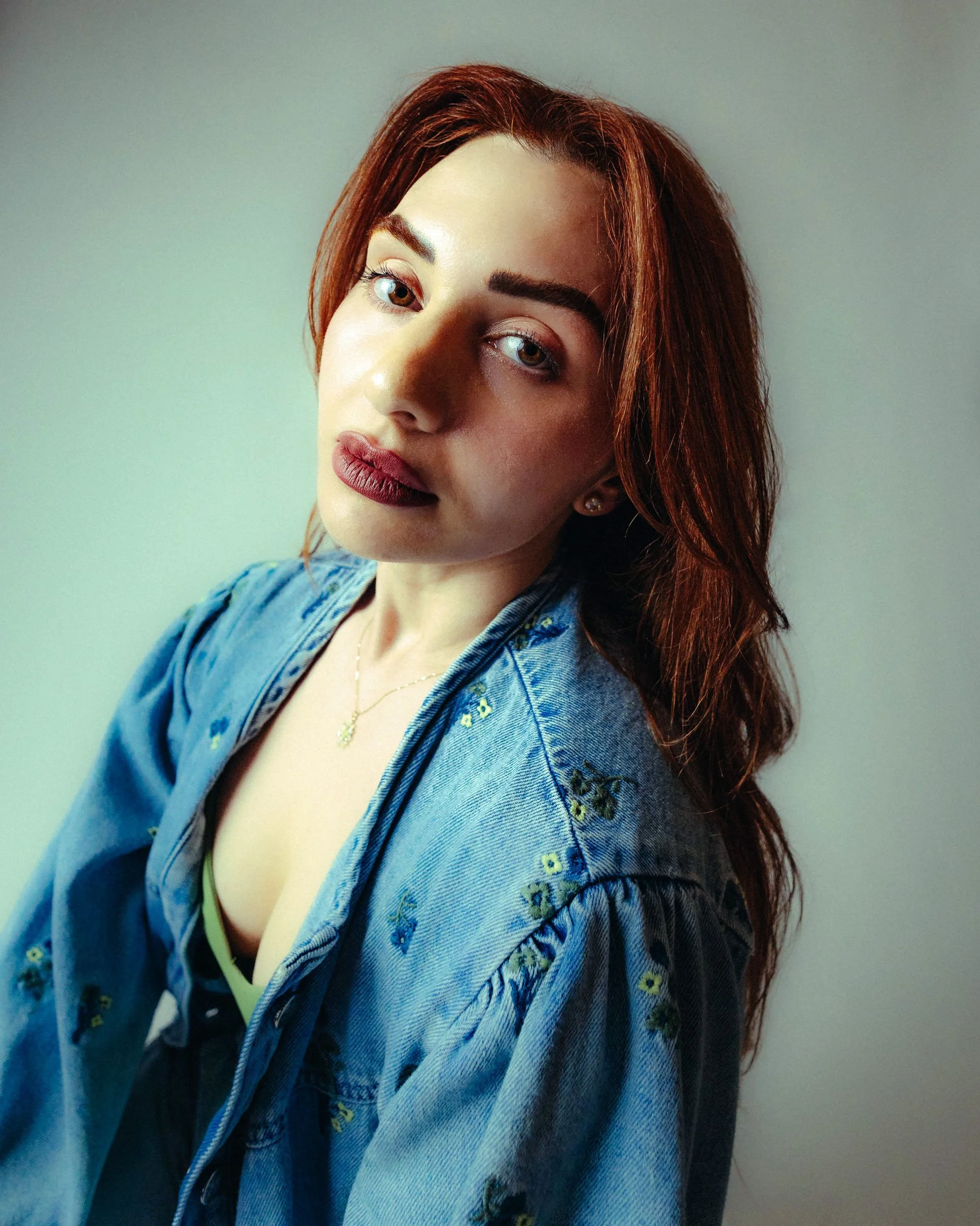 Close-up of a young woman with red hair, green eyes, and makeup, wearing a denim jacket and a necklace.