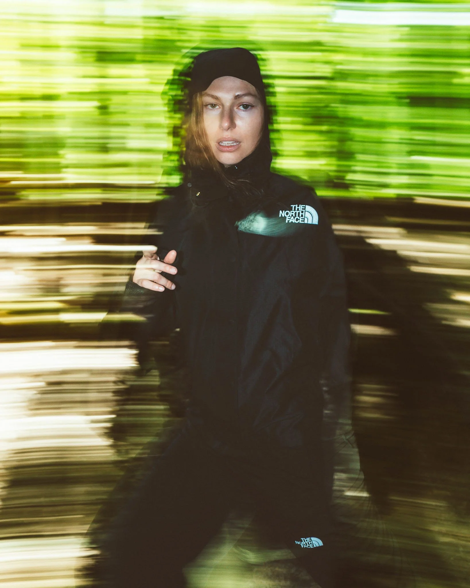 A woman with long brown hair, wearing a black beanie and a black The North Face jacket, standing in front of a blurred background of green trees. The photo has motion blur, creating a sense of movement.