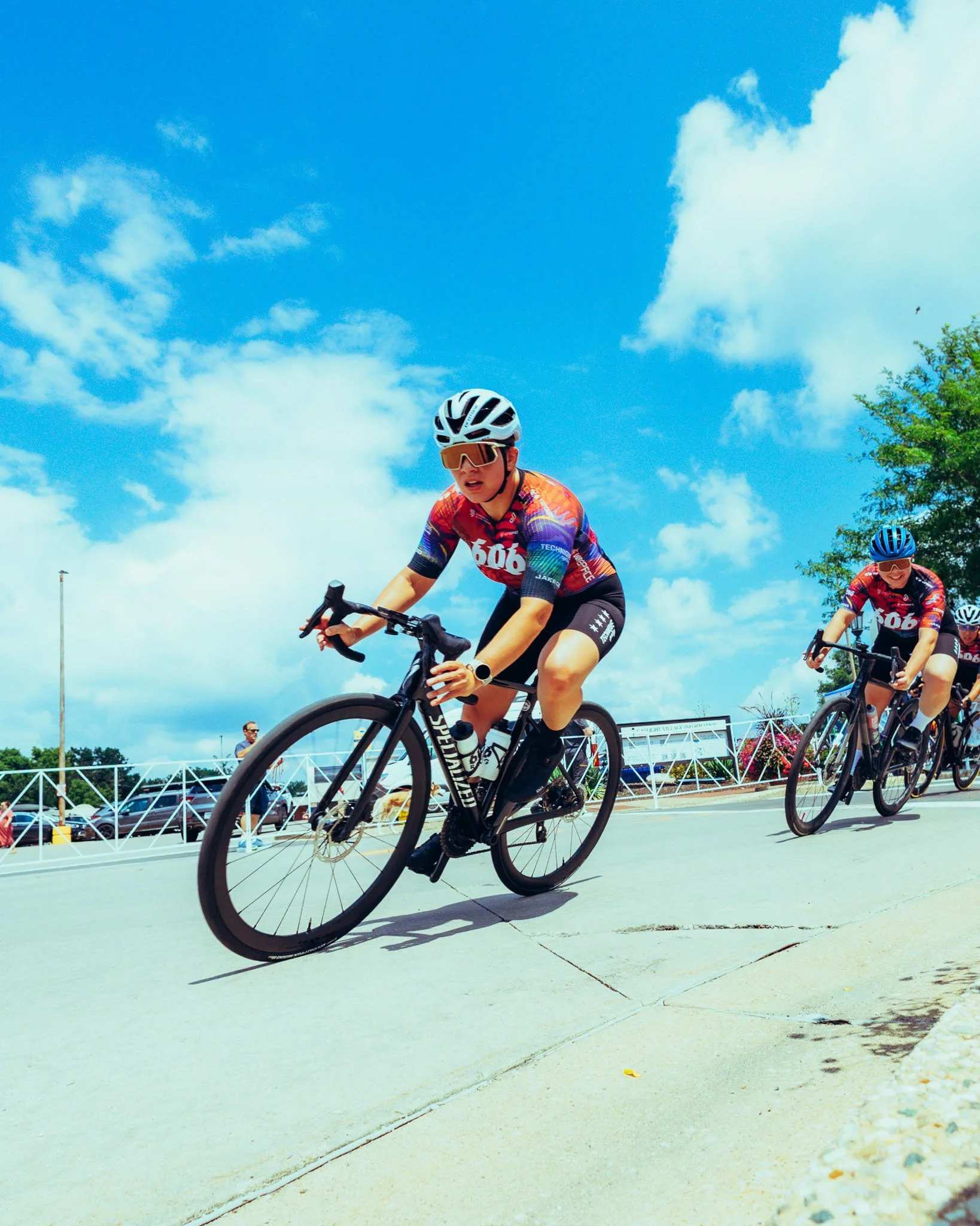 Cyclists racing outdoors under a clear, partly cloudy sky with trees and a parking lot in the background.