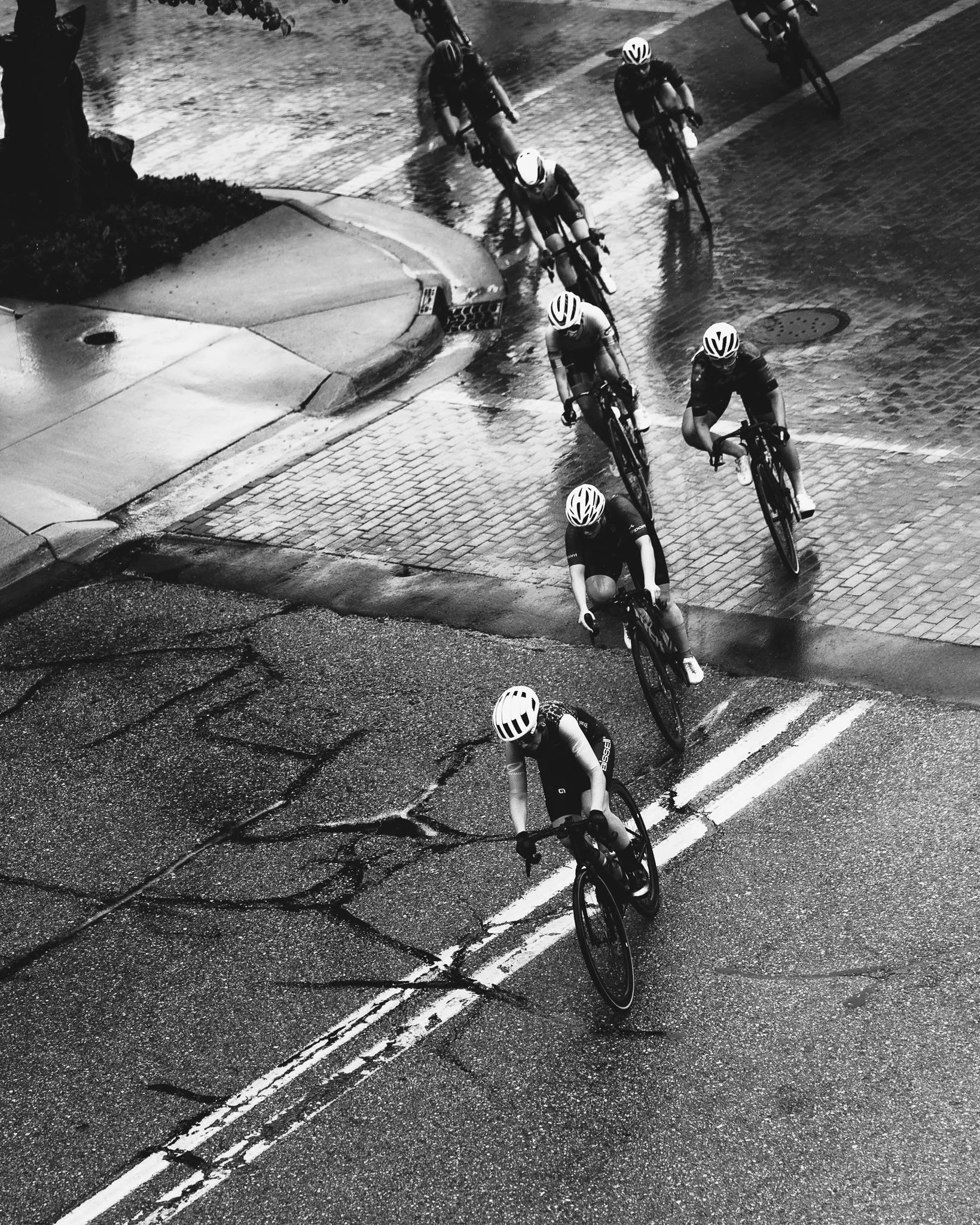 A group of cyclists riding on a wet city street at night, with some reflecting light from puddles and streetlights, captured from an overhead perspective.