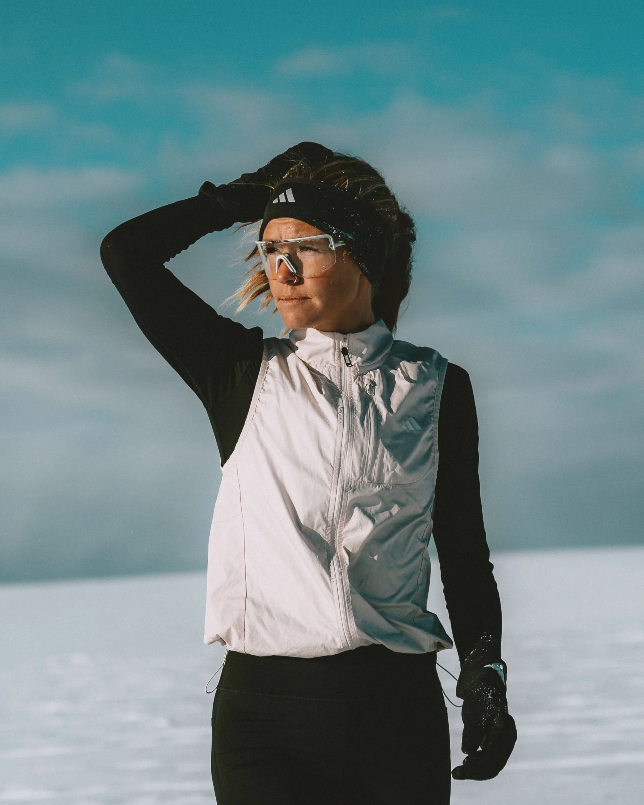A woman dressed in sports gear, wearing a white jacket, black gloves, a black headband, and transparent sports glasses, standing outdoors against a cloudy sky.