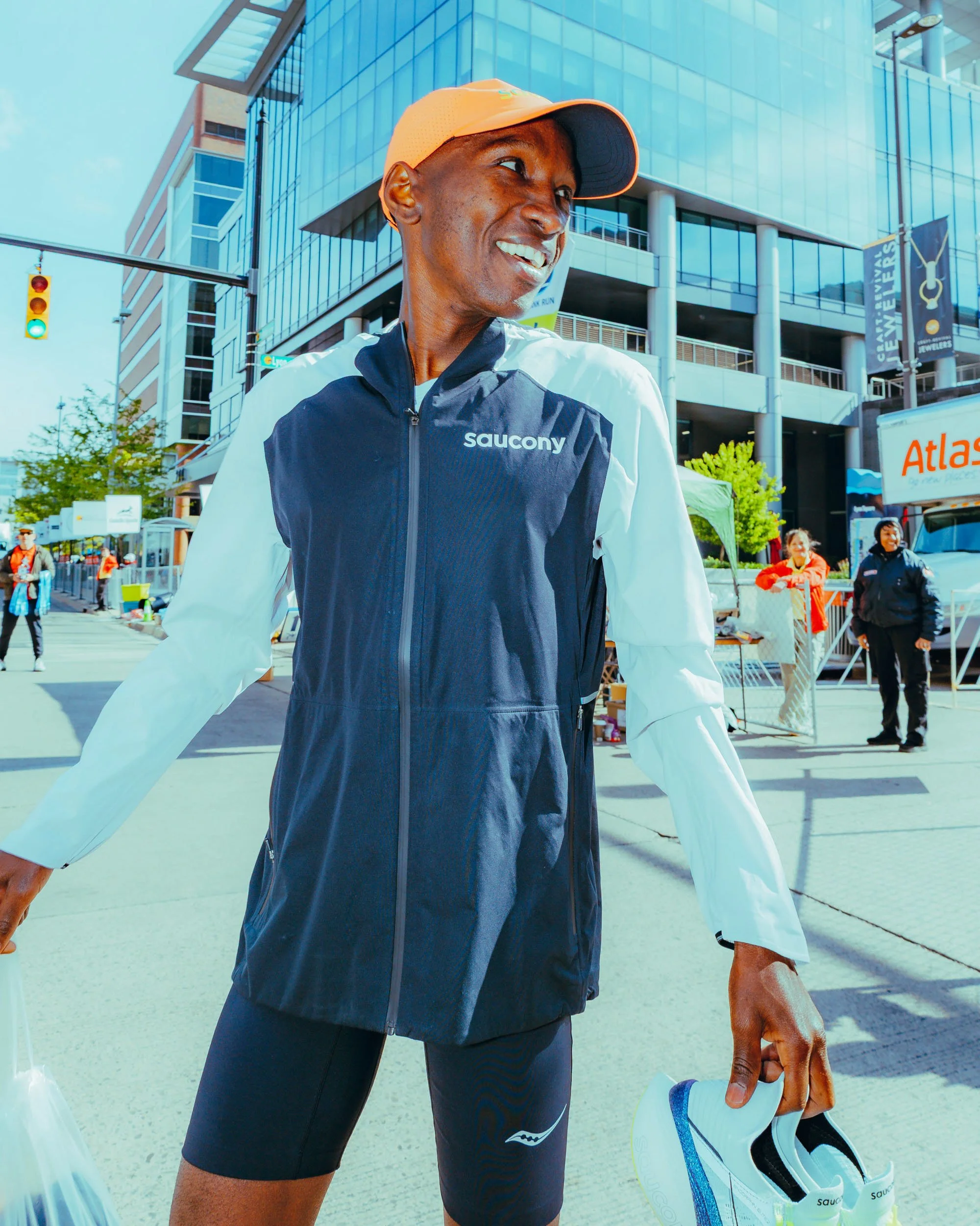 A man in athletic clothing, including a Saucony branded jacket and shorts, is holding a pair of white running shoes with blue accents, and wearing an orange cap. He is outdoors in an urban area with modern glass buildings, traffic lights, and other p
