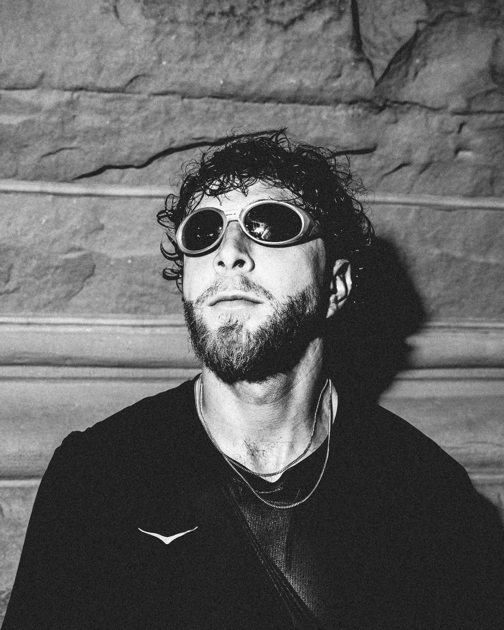 A person with curly hair, a beard, and wearing round sunglasses and a black shirt, looking at the camera against a textured wall background.