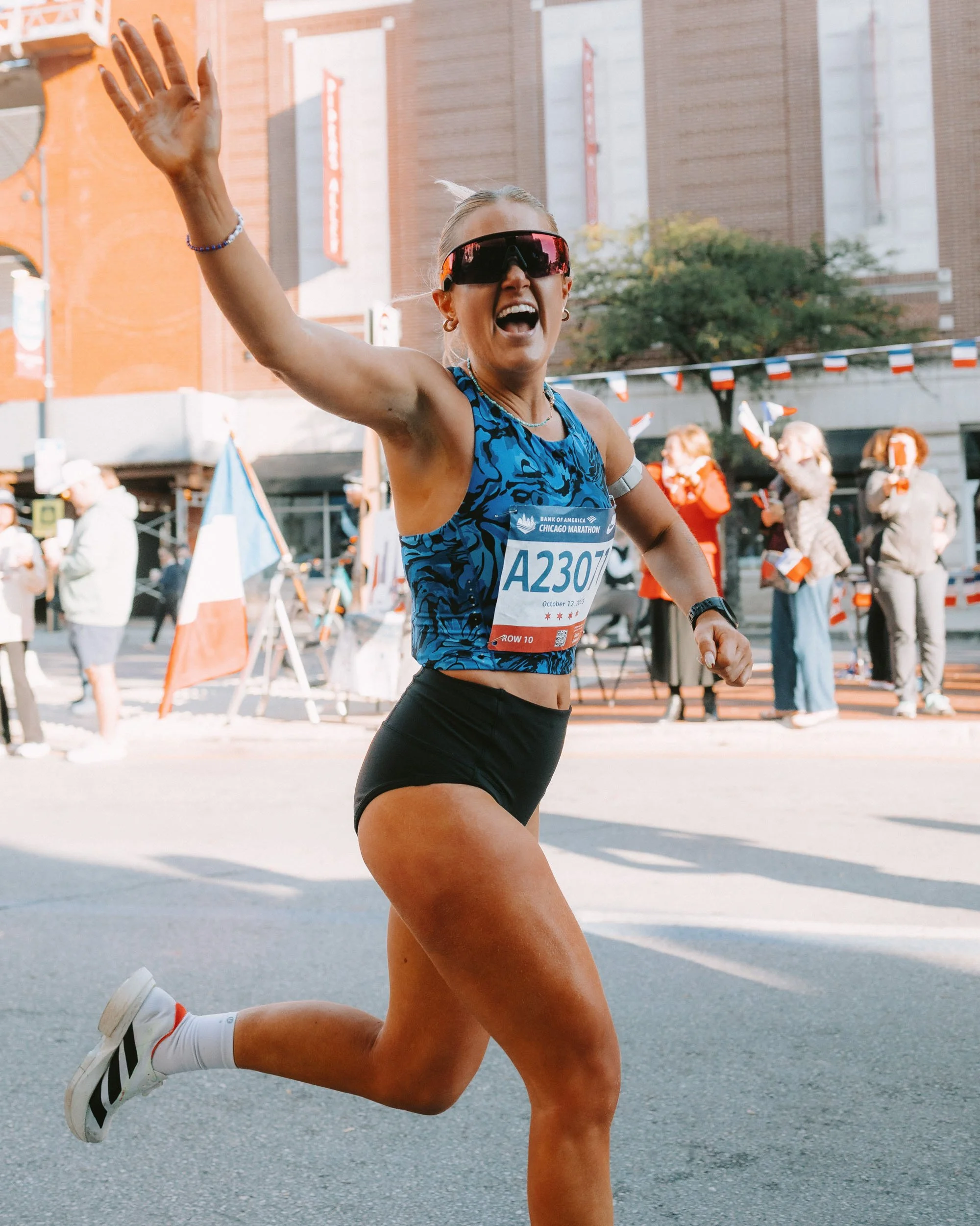 A woman running in a race, wearing sunglasses and athletic attire, with a race bib number A2307, on a city street with spectators in the background.