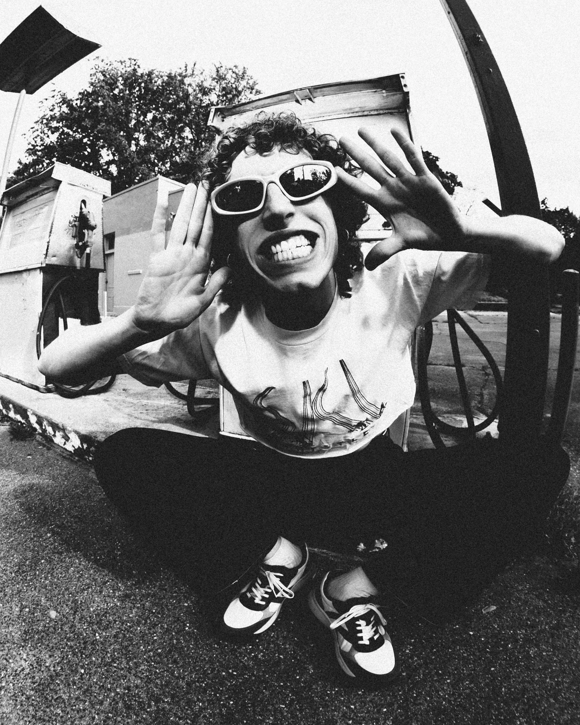 Black and white photograph of a young person with curly hair wearing sunglasses and grinning, sitting outdoors with trees and structures in the background, and their legs crossed in sneakers.
