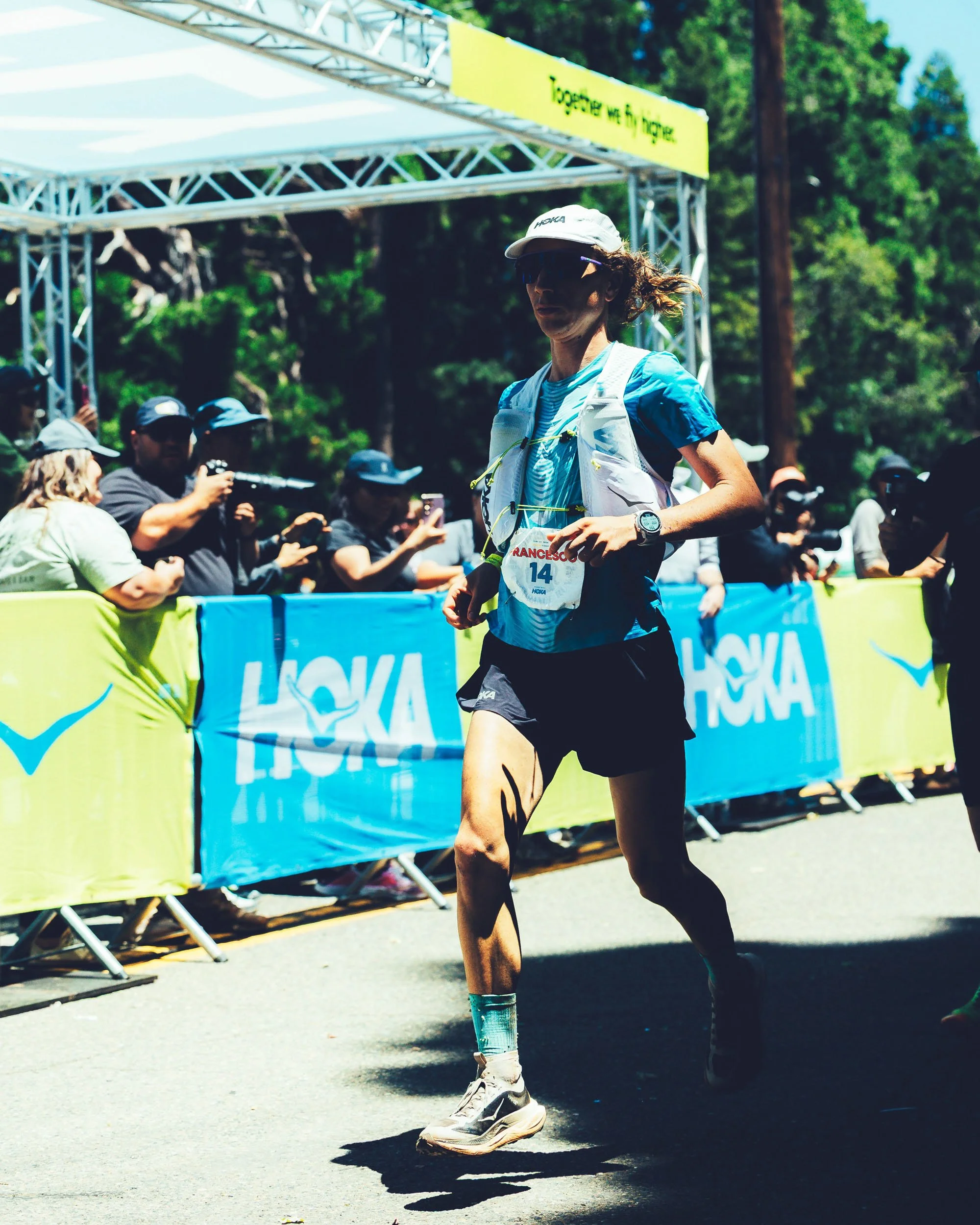 A woman running in a race, wearing a blue and white athletic outfit, a white cap, sunglasses, and a race bib numbered 14, with a crowd and banners in the background.