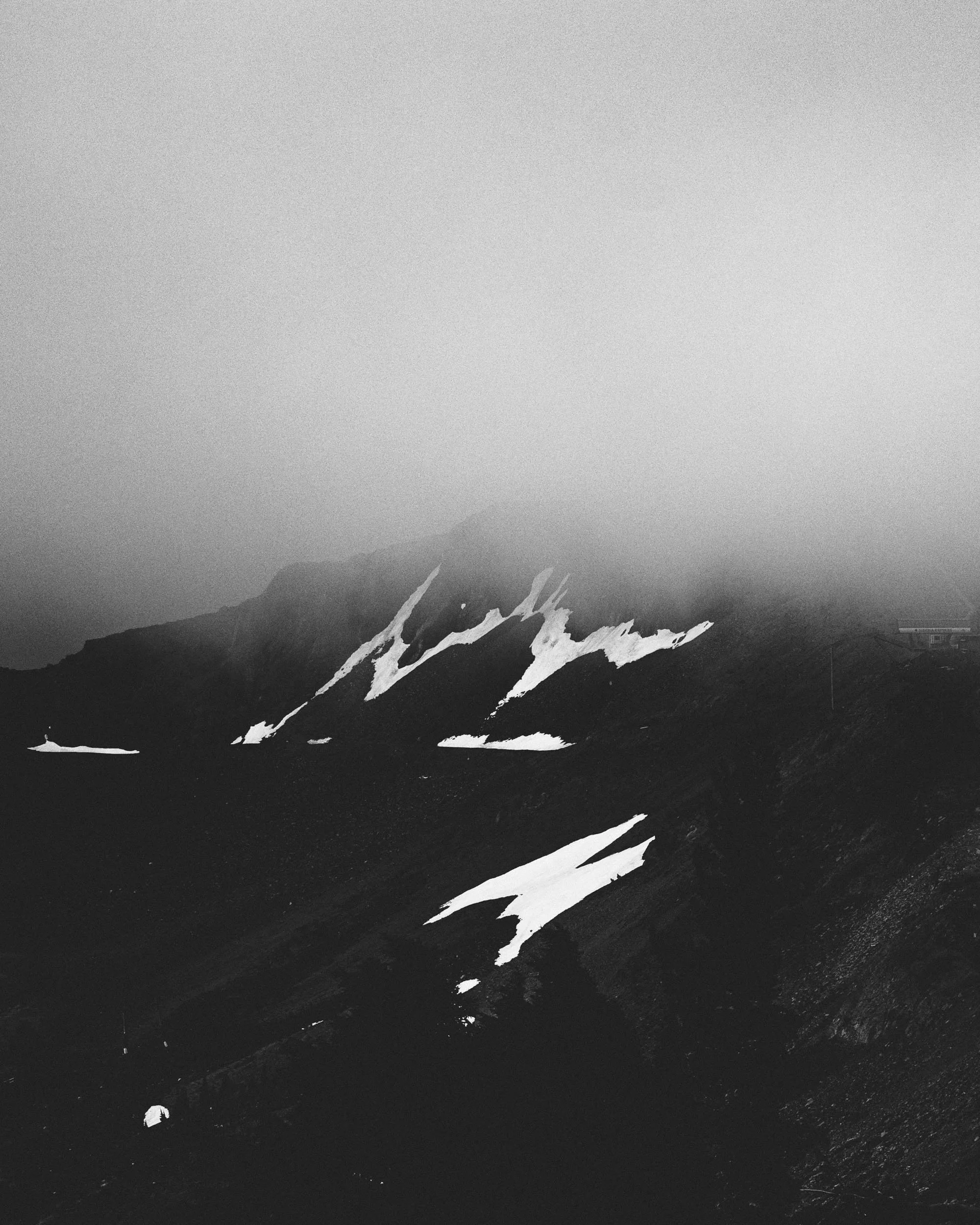 A black and white photo of snow-capped mountains under a cloudy sky with land in the foreground.