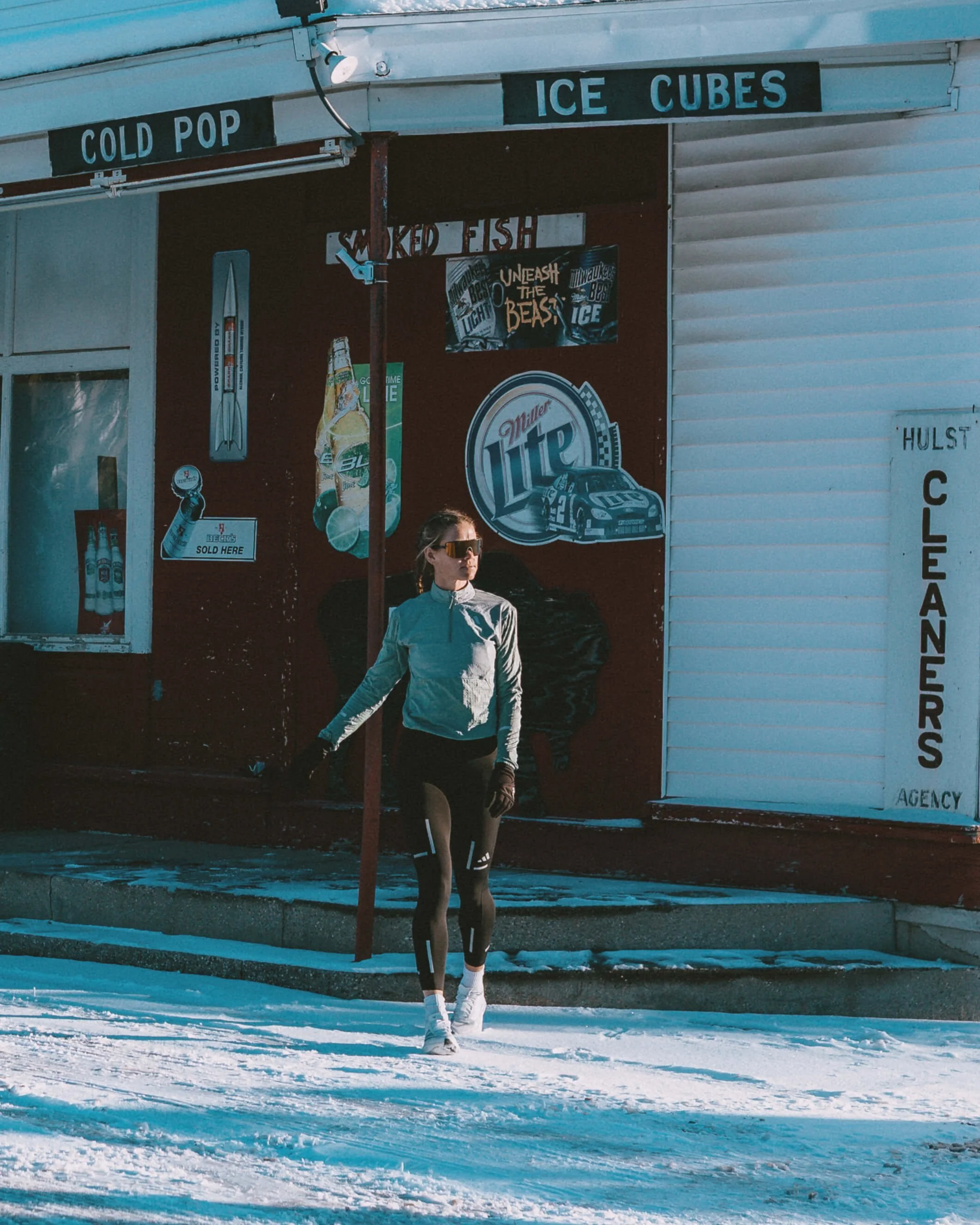 A woman standing in front of a storefront selling cold pop, ice cubes, and smoked fish, with snow on the ground. She is wearing sunglasses, a light jacket, leggings, and sneakers, and appears to be looking to her left.