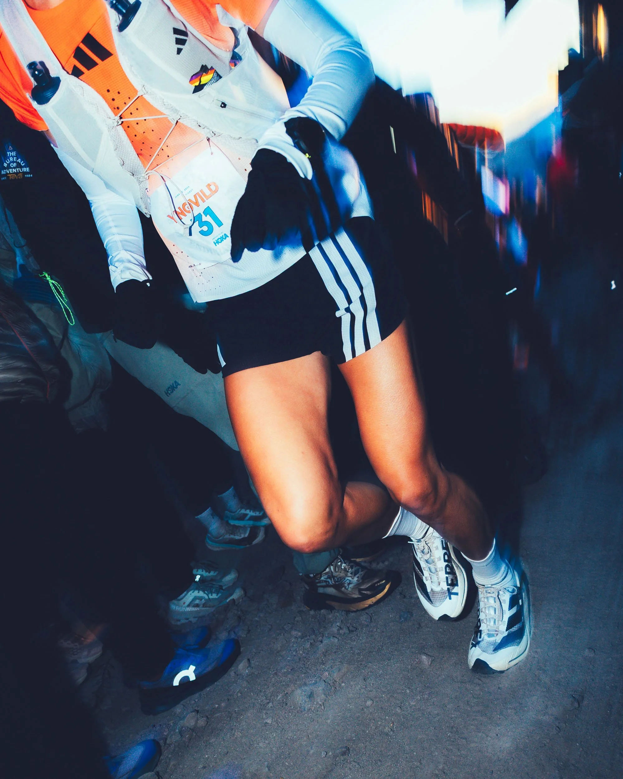 Close-up of people’s legs and feet, wearing sports shoes, sitting on a rocky outdoor surface with a blurry background.