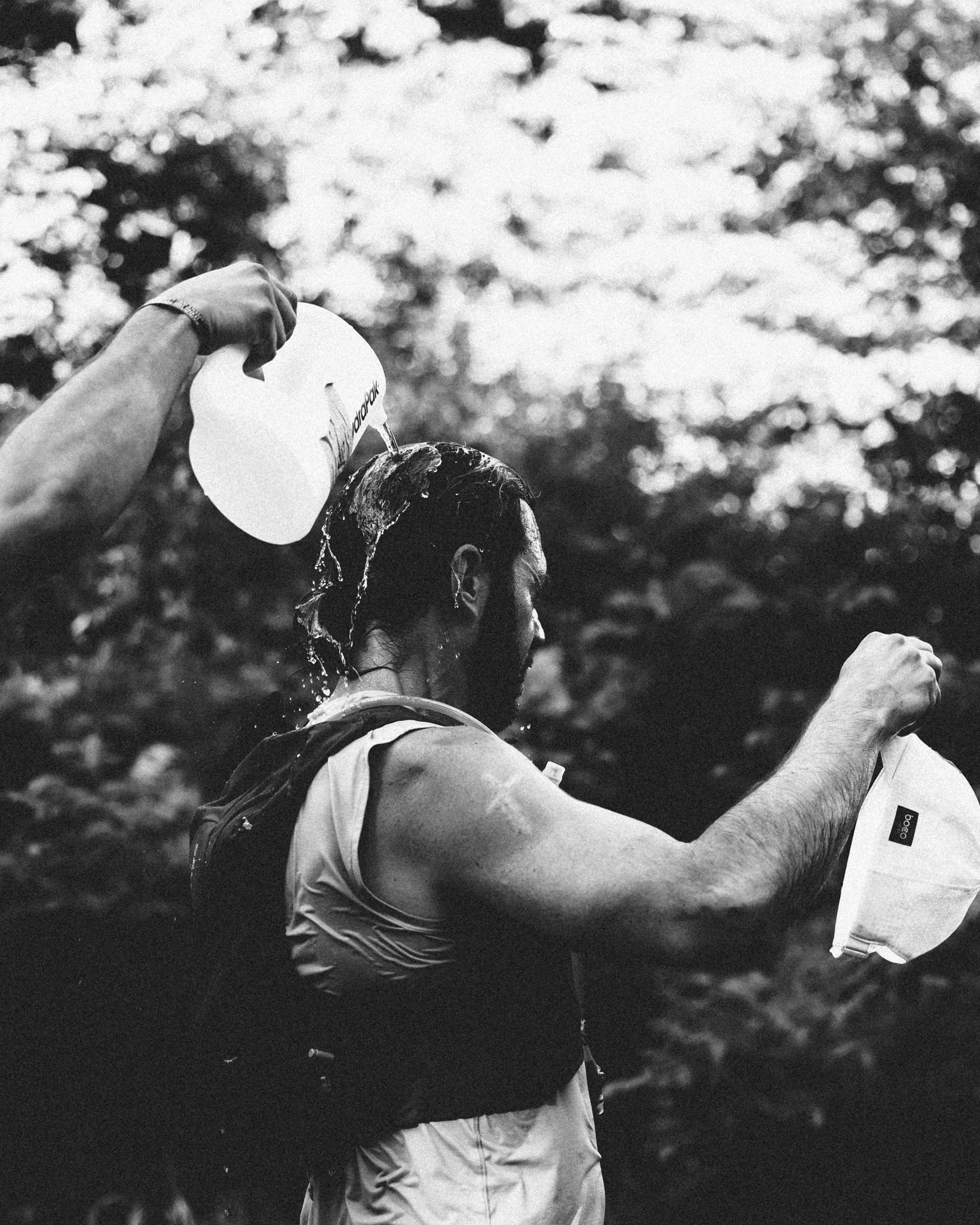 A person pouring water over another person's head outdoors with trees blurred in the background.