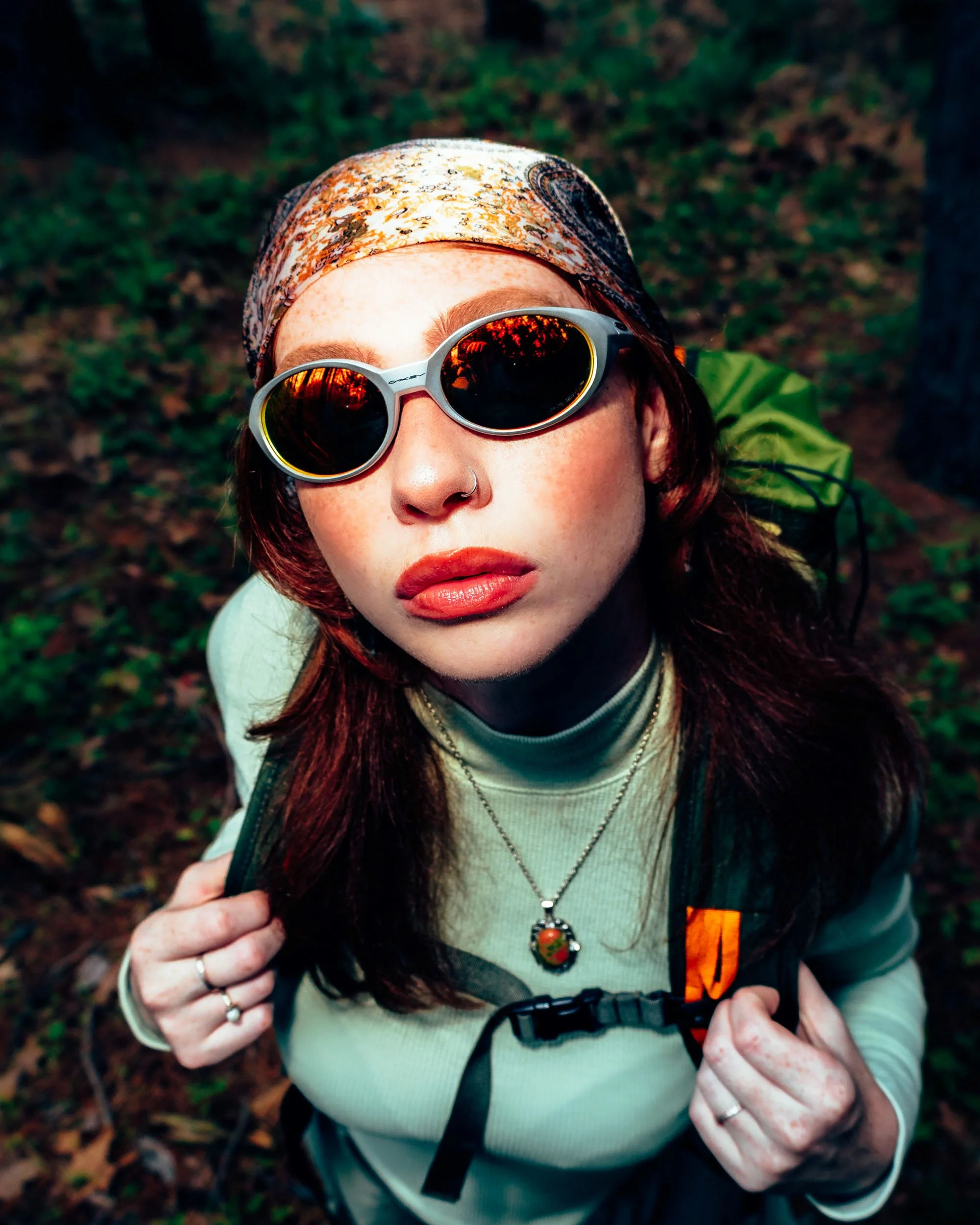 A woman wearing sunglasses and a colorful bandana on her head, outdoors in a wooded area, holding a camera strap with both hands.