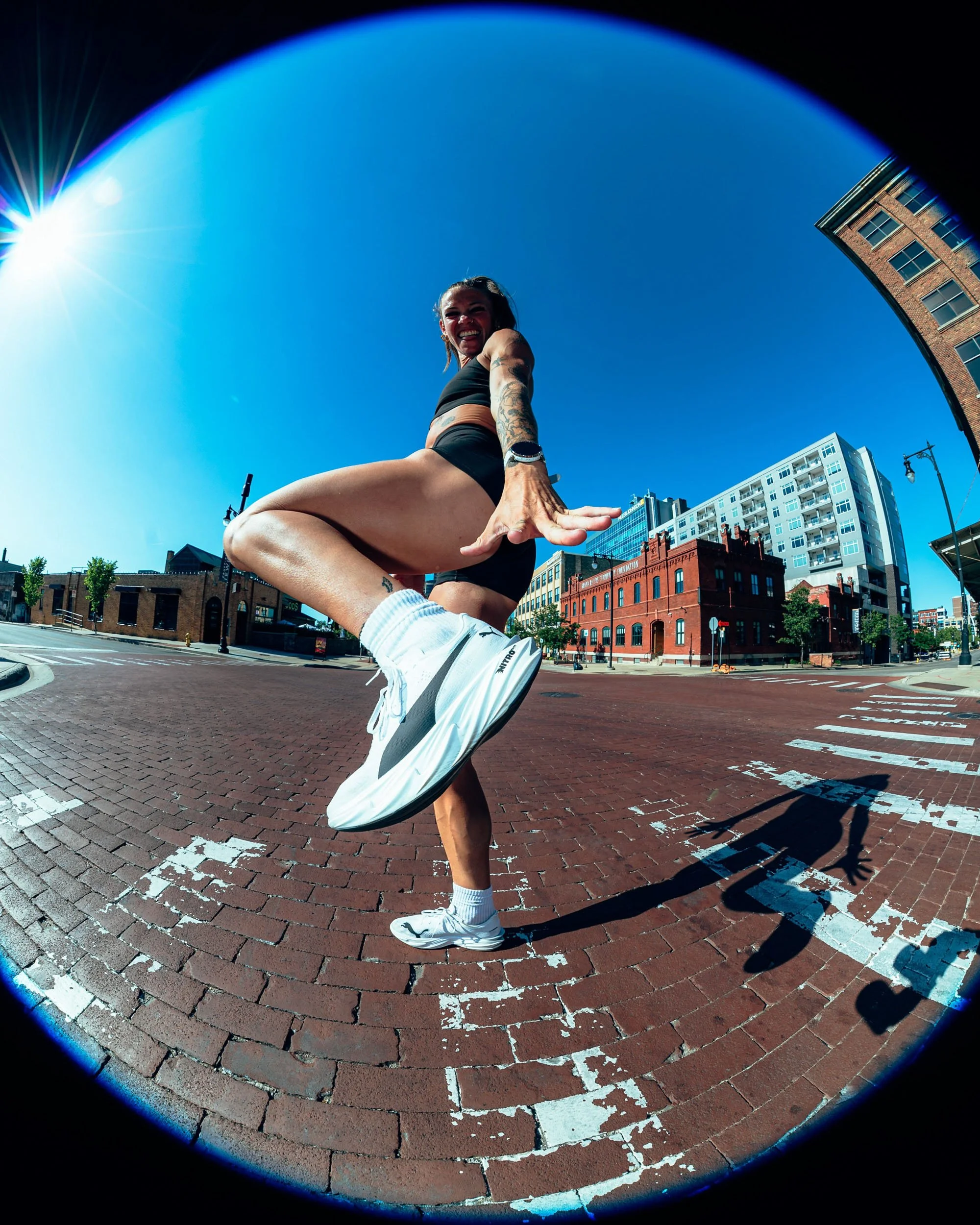 A woman with tattoos on her arm, wearing a black sports bra and shorts, performing a dance or yoga pose on a city street with a brick crosswalk, surrounded by tall buildings and a clear blue sky.