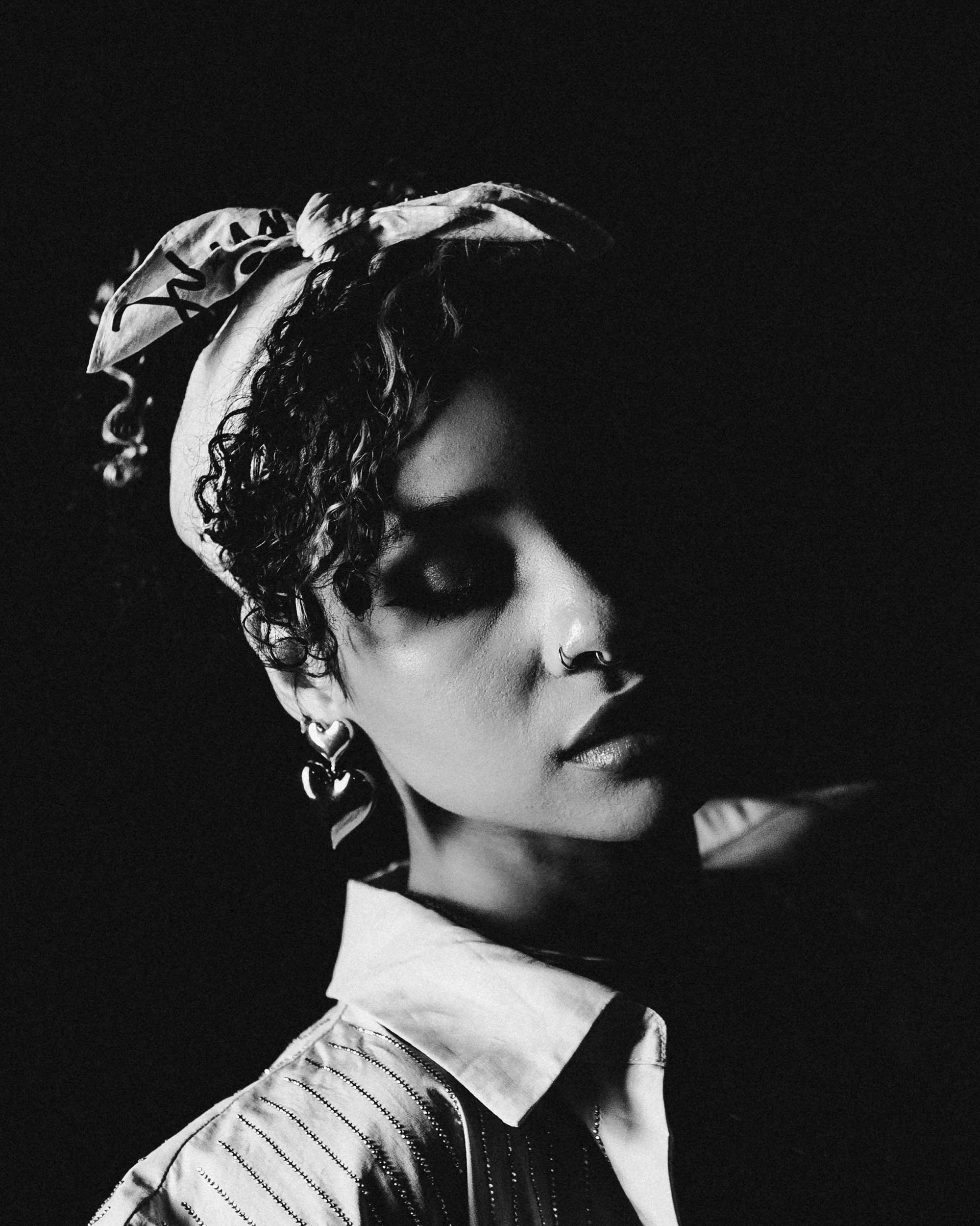Close-up black and white photo of a woman with curly hair and hoop earrings, wearing a patterned jacket and a white shirt, with her eyes closed against a dark background.