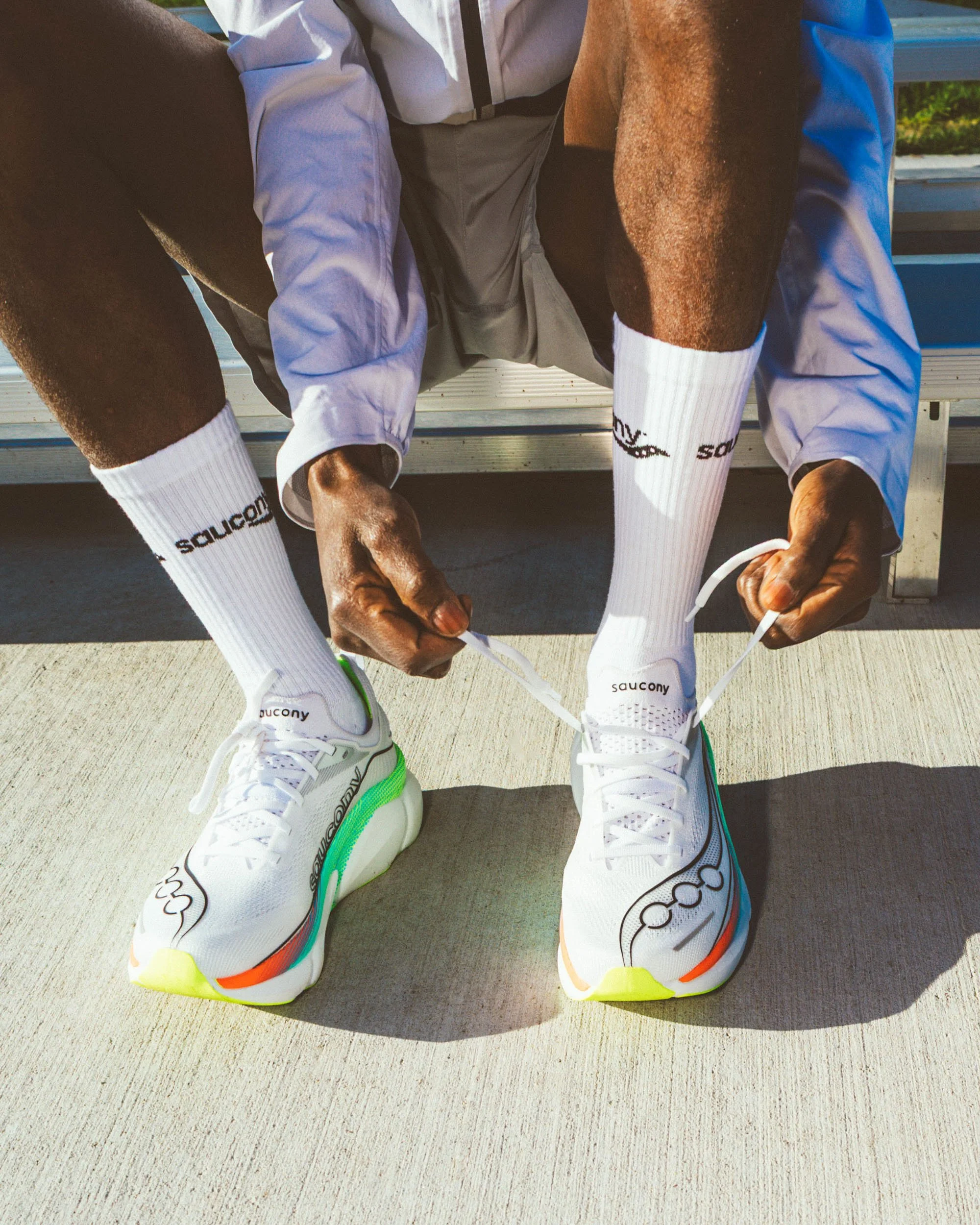 Person in white athletic shoes with neon accents tying their shoelaces while sitting on a bench, wearing white socks with black text, white pants, and a blue and gray jacket, with another person's legs visible on each side.