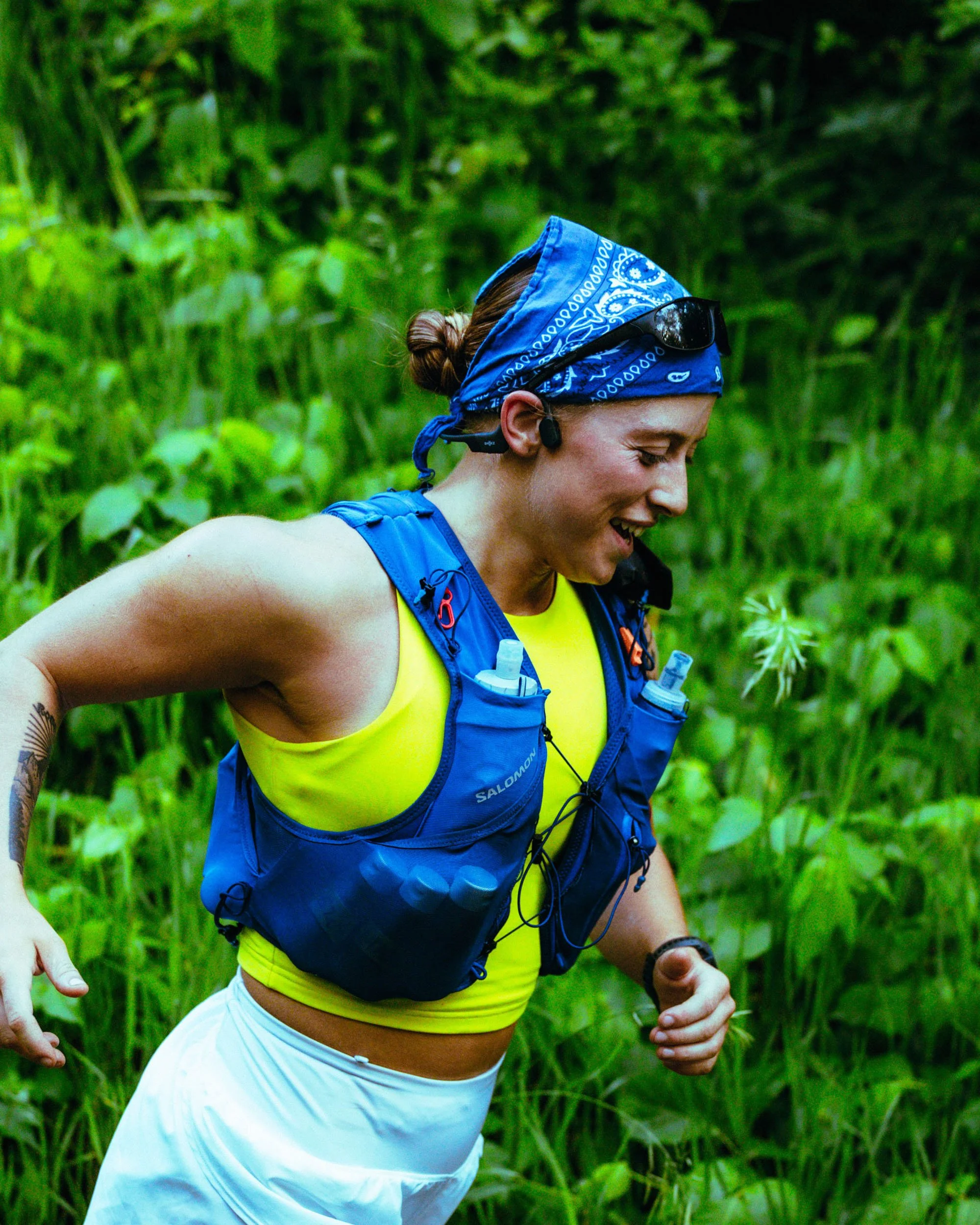 A woman running through a lush, green outdoor setting. She is wearing a blue bandana, sunglasses on her head, a mint green tank top, a blue hydration pack, and white shorts.