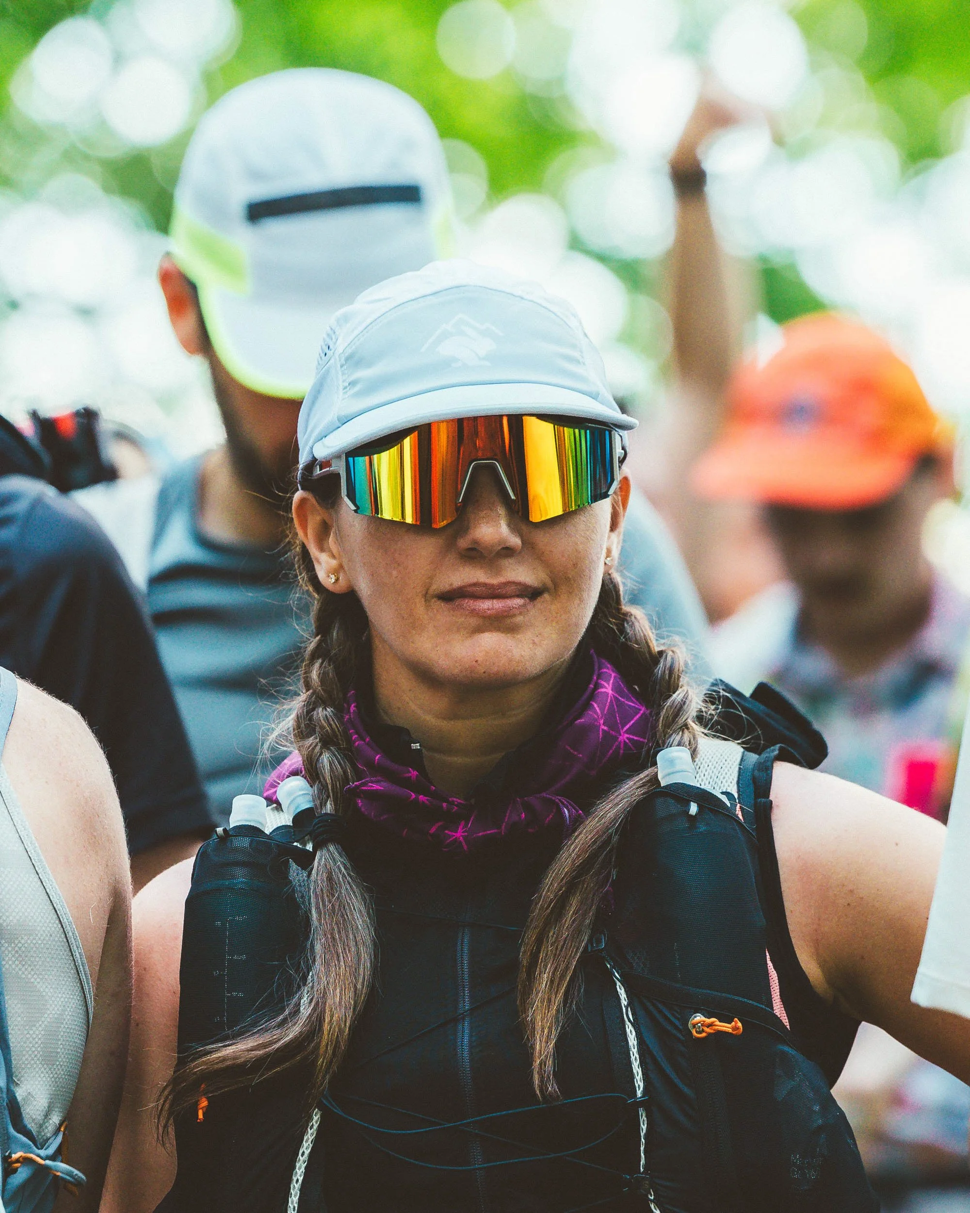 Close-up of a woman wearing reflective sports sunglasses, a light blue cap, and a purple neck gaiter, with braided hair, during an outdoor activity with other people in the background.