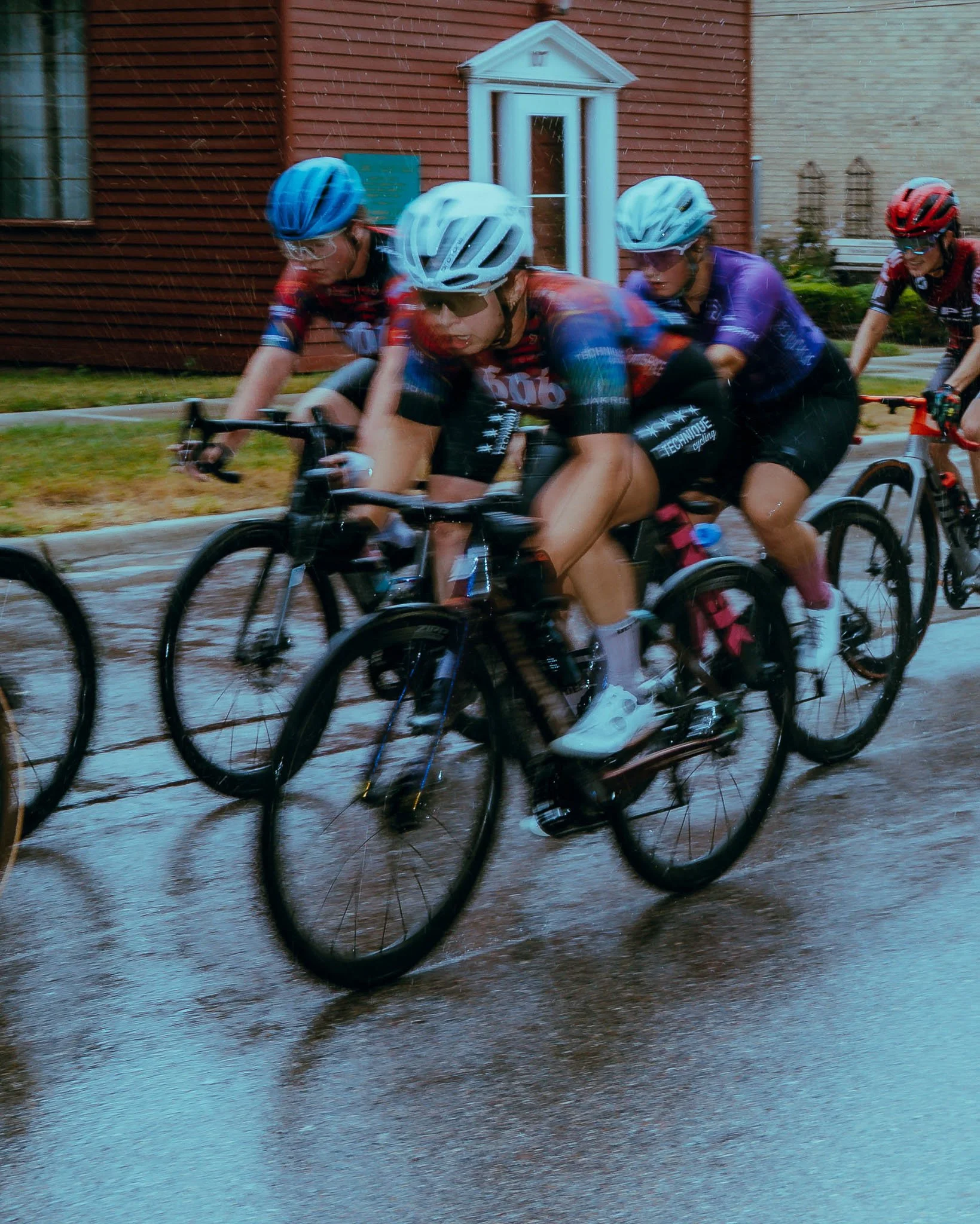 Cyclists racing through a rain-soaked street, all wearing helmets and cycling gear, with blurred background of houses.