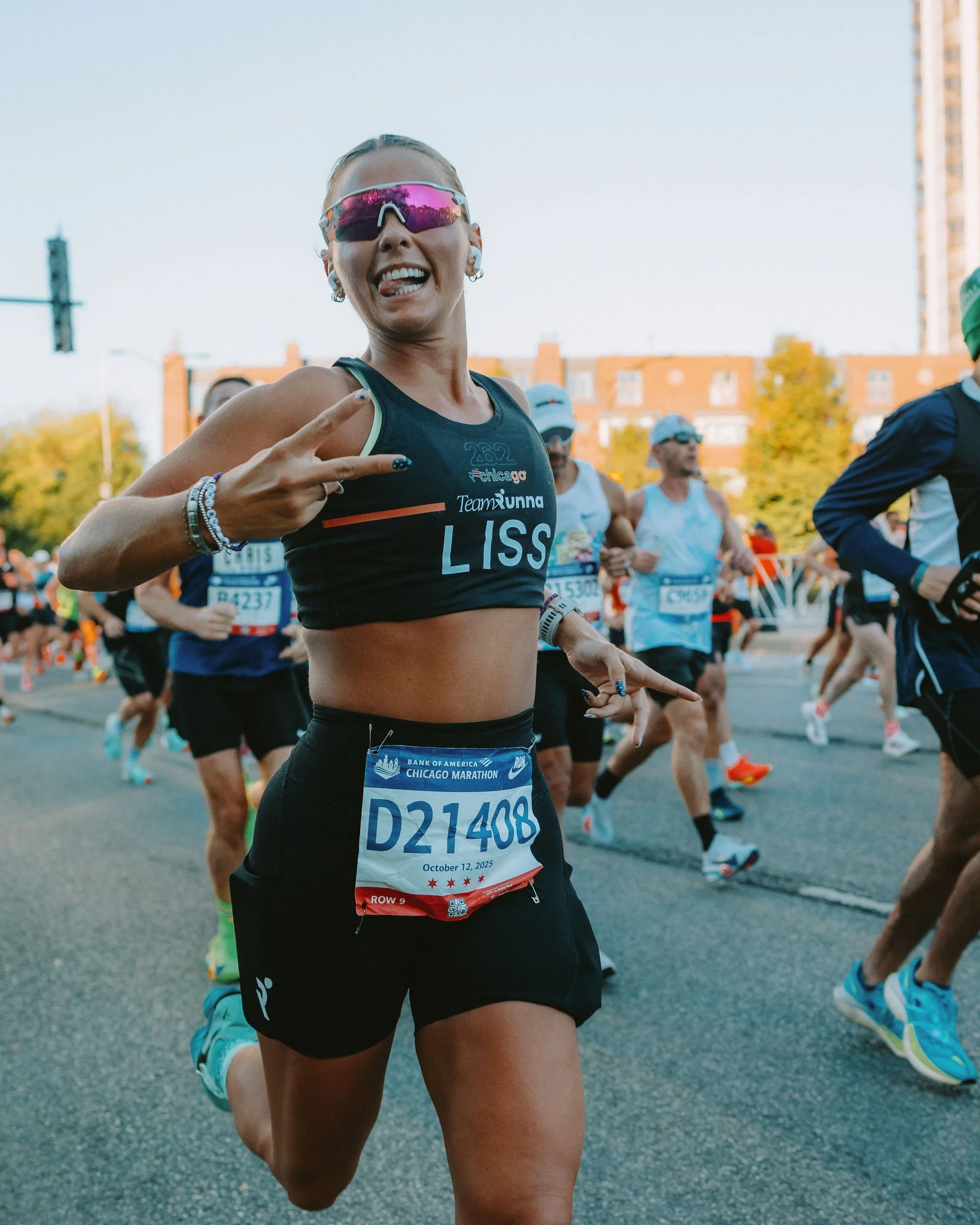 A female marathon runner making a victory sign with her fingers as she runs in a race, wearing sunglasses, a black sports bra, and black shorts. Other runners are visible in the background on a city street with buildings and trees.