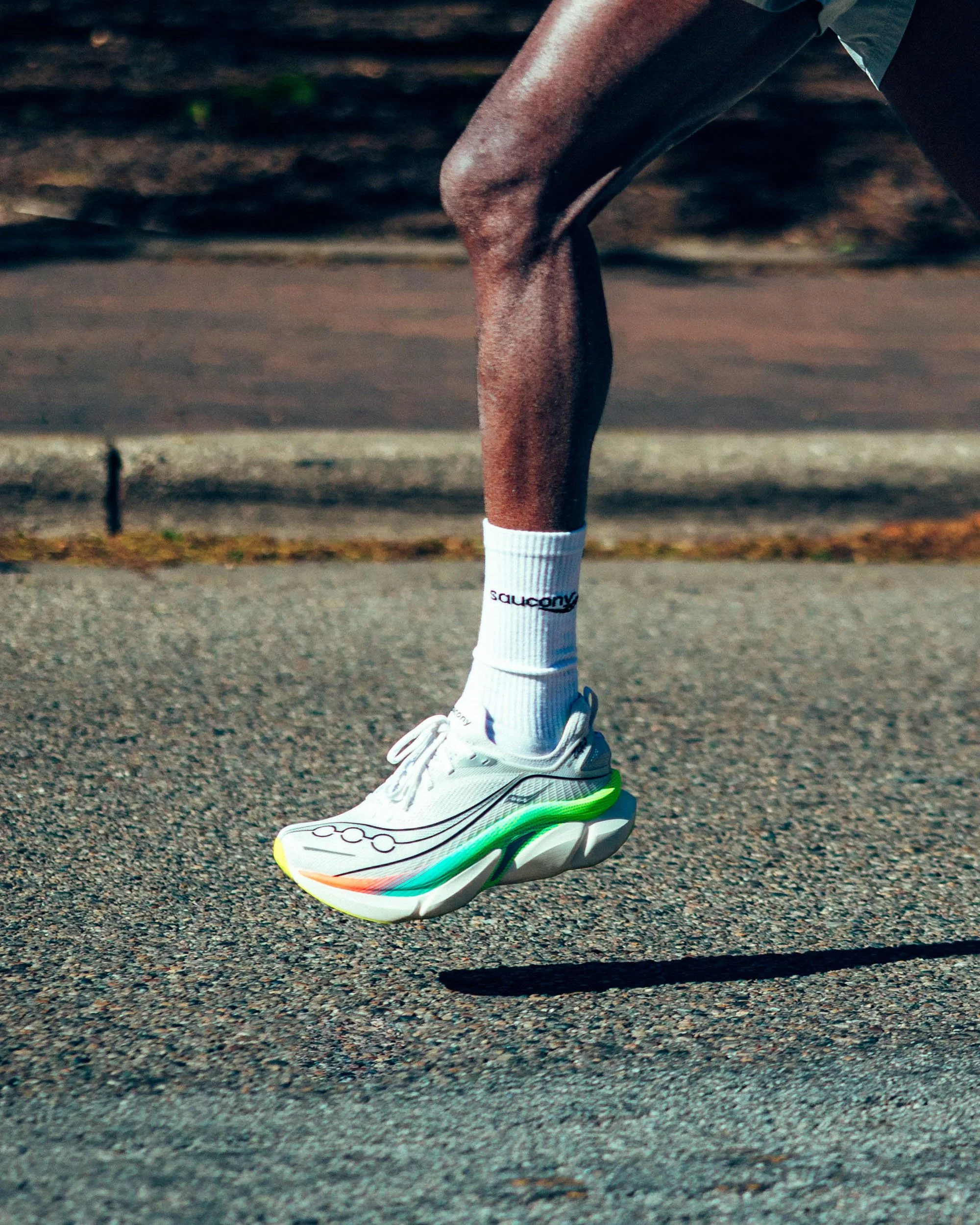 Close-up of a person running, focusing on their right leg, wearing a white running shoe with neon green and orange accents, a white sock, and dark shorts, on an asphalt pavement.