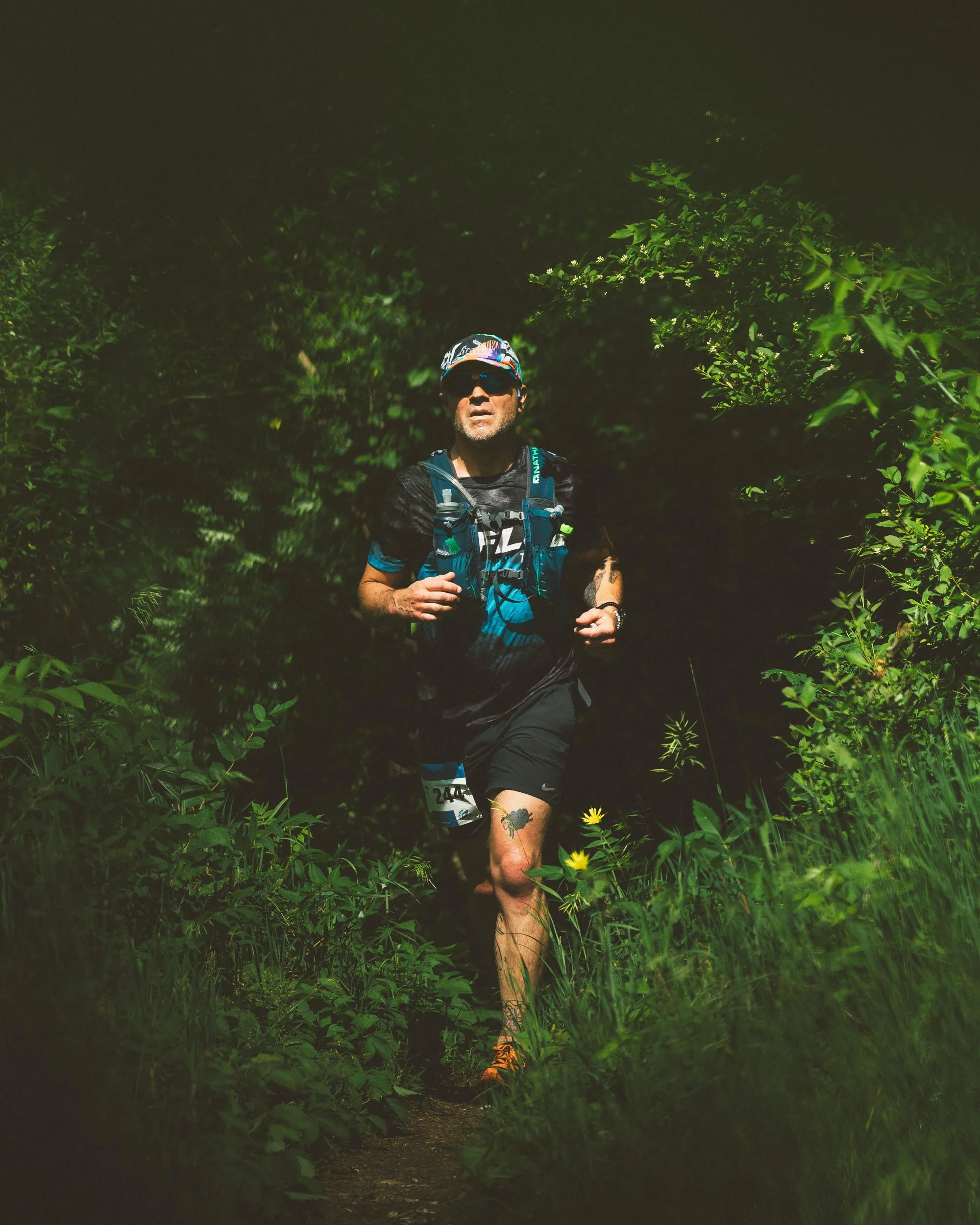 A man running on a trail through dense green foliage at night, wearing a black sports shirt, a hydration vest, a colorful cap, sunglasses, and orange trail running shoes.