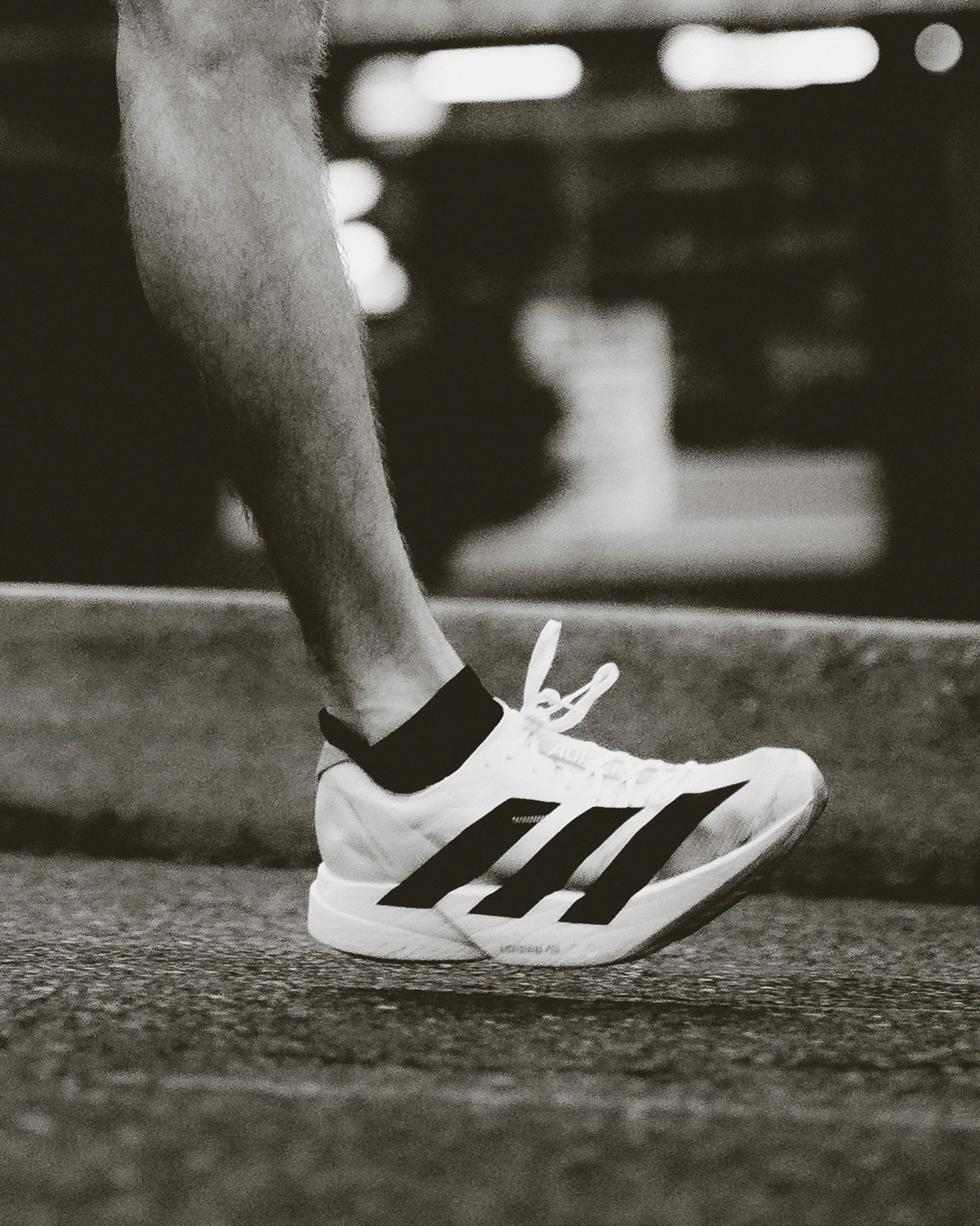Close-up of a person's foot wearing a white sneaker with black stripes, black ankle sock, and dark shorts, standing on wooden flooring. Background is blurred.
