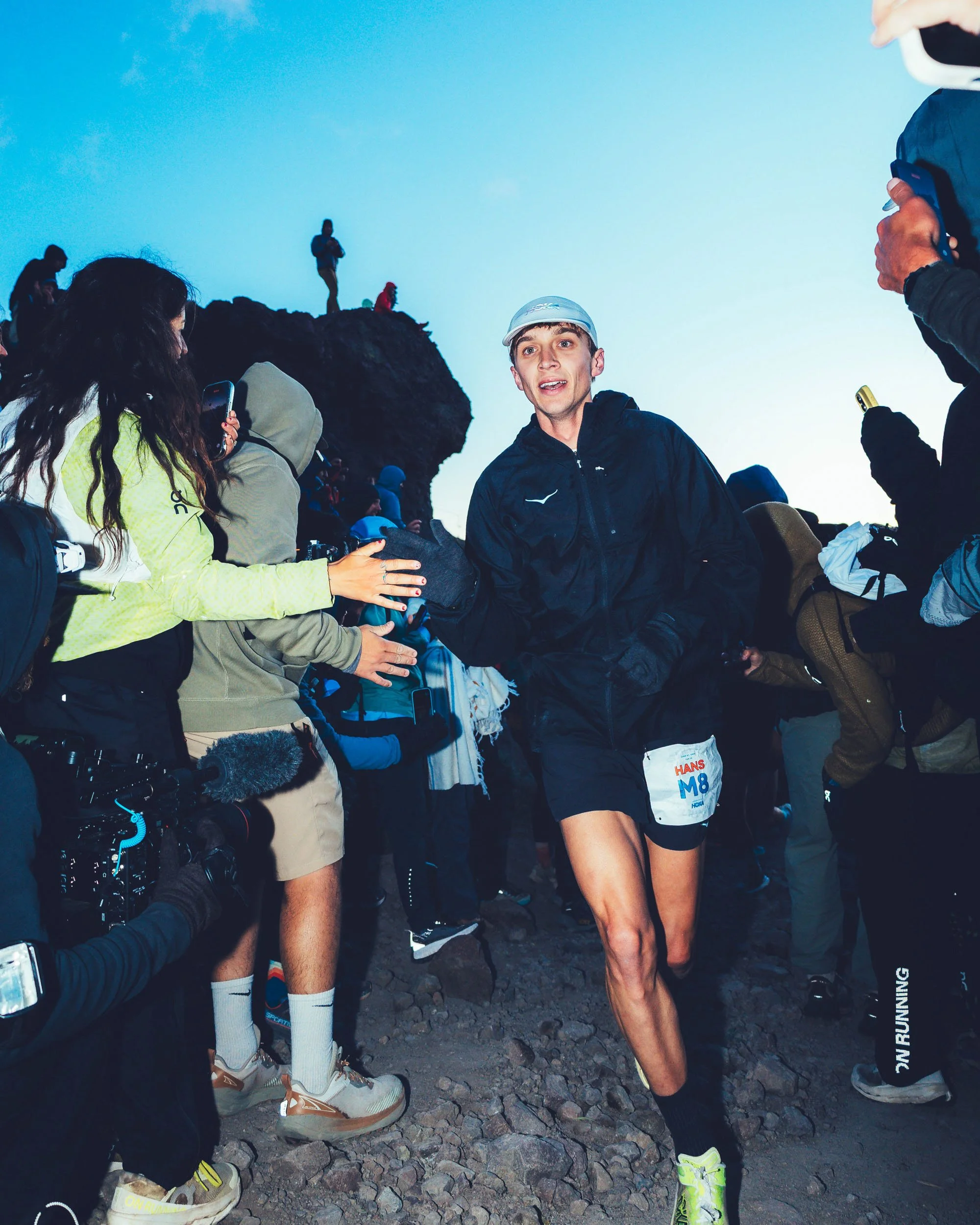 A male runner wearing a black jacket, black shorts, neon yellow running shoes, and a white cap is talking to a woman with long dark hair, wearing a yellow and gray jacket and beige shorts, at a mountainous outdoor location with rocks and other people