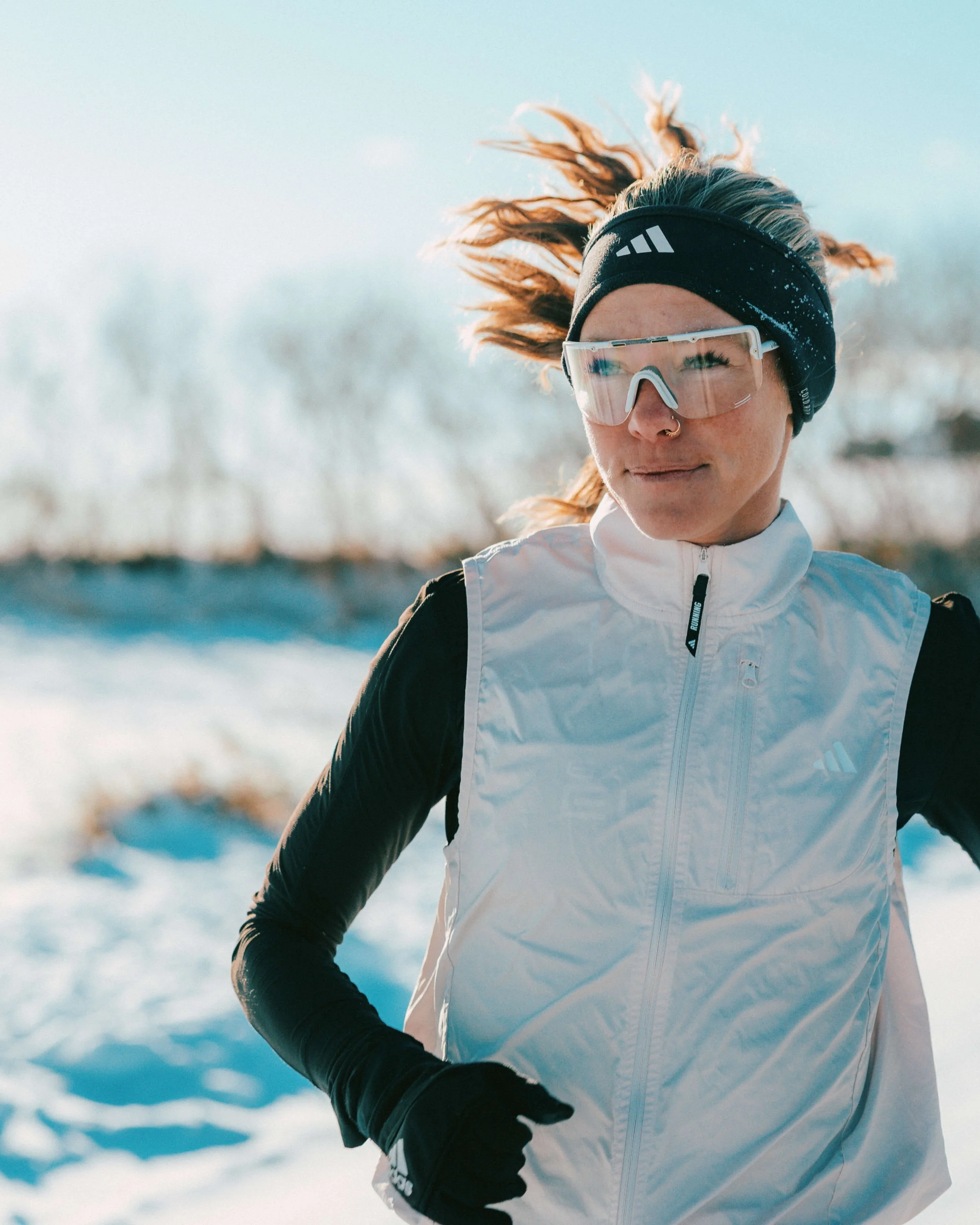 A woman with curly hair wearing a black headband, sunglasses, a white jacket, and black gloves, running outdoors in a snowy landscape.